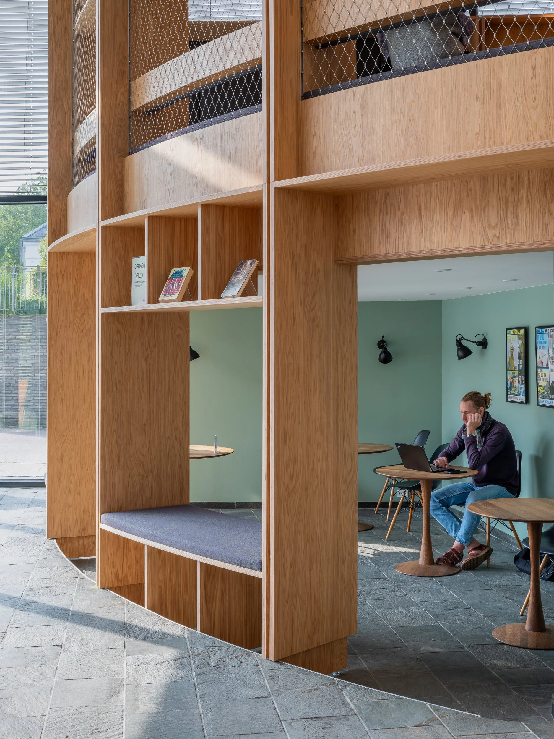 Interior of a modern cafe or lounge area with a wooden partition that includes open shelving and a built-in seat. A man is sitting at a small round table, working on a laptop, with framed art on the mint green wall behind him.