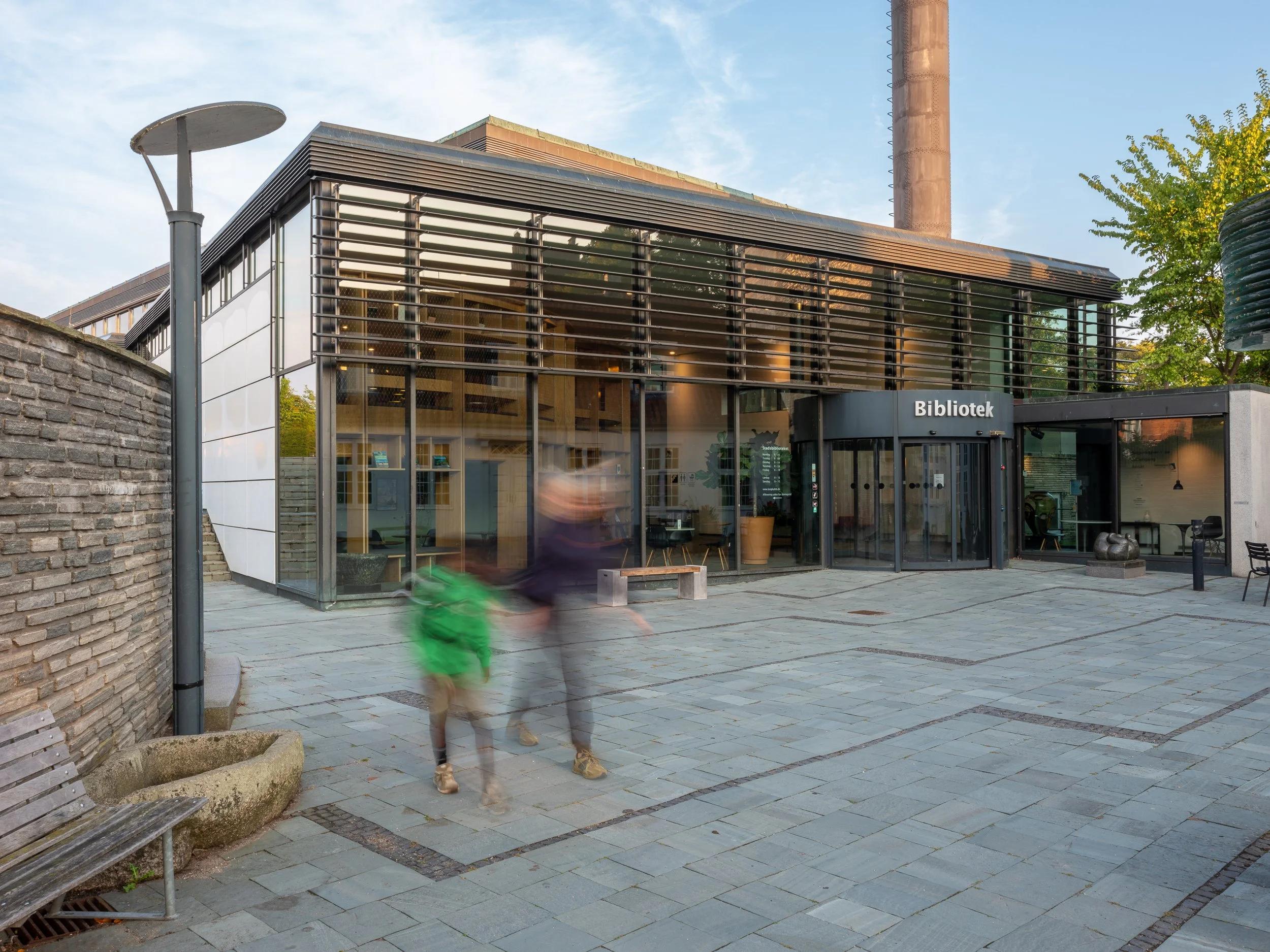 Modern library building with glass and wood facade, labeled 'Bibliotek,' on a paved outdoor area with a bench, a lamppost, and some blurred people walking.