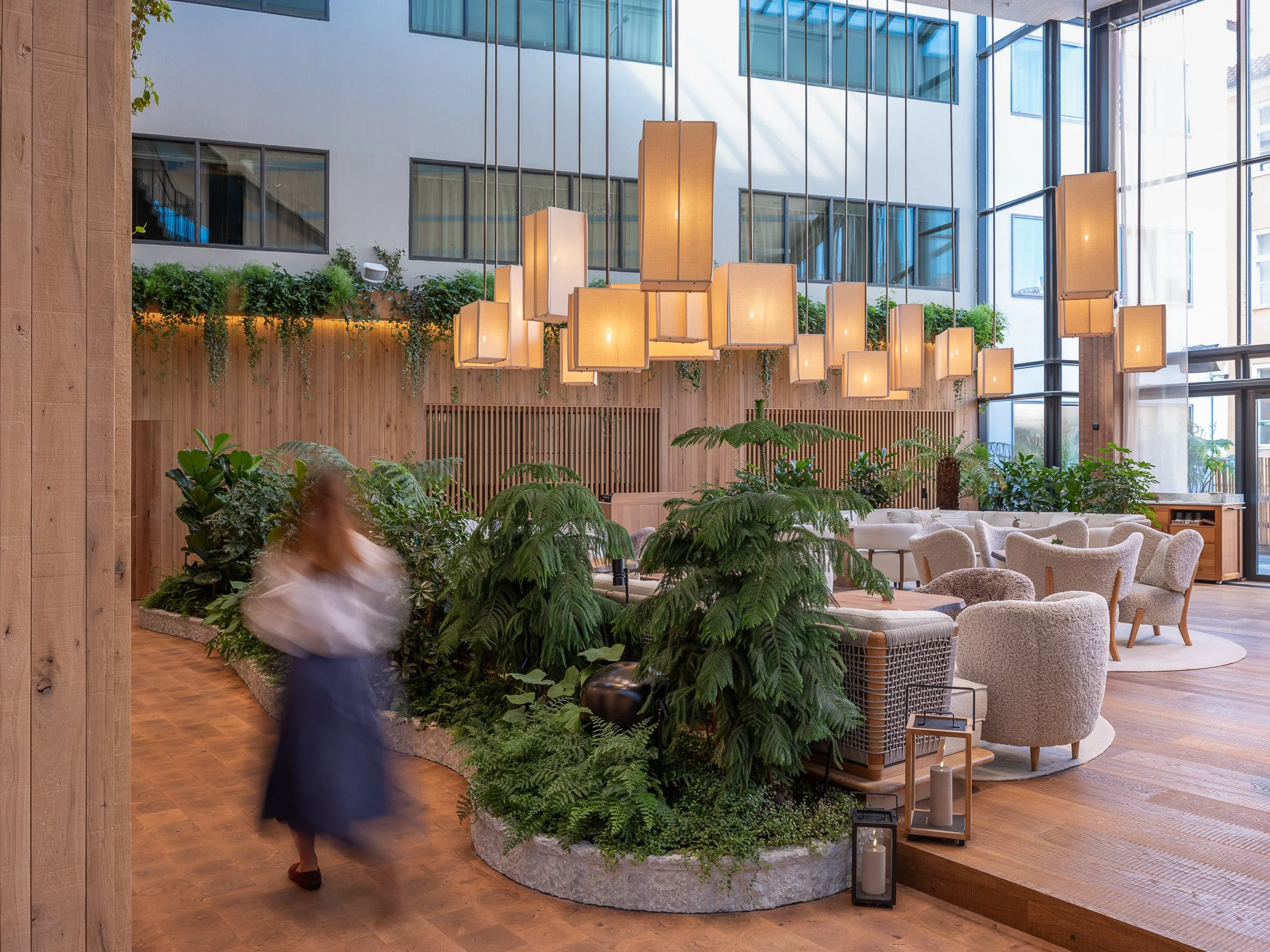 Interior of a modern lobby or lounge area with wooden floors, lush greenery, beige and white furniture, and large hanging light fixtures, featuring a glass wall allowing natural light.