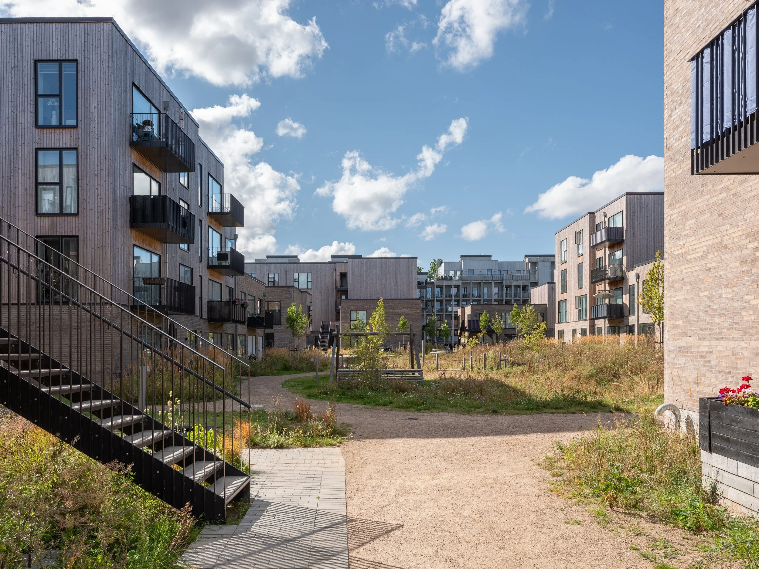Modern residential apartment complex with multiple buildings, green outdoor space, and walking paths under a partly cloudy sky.