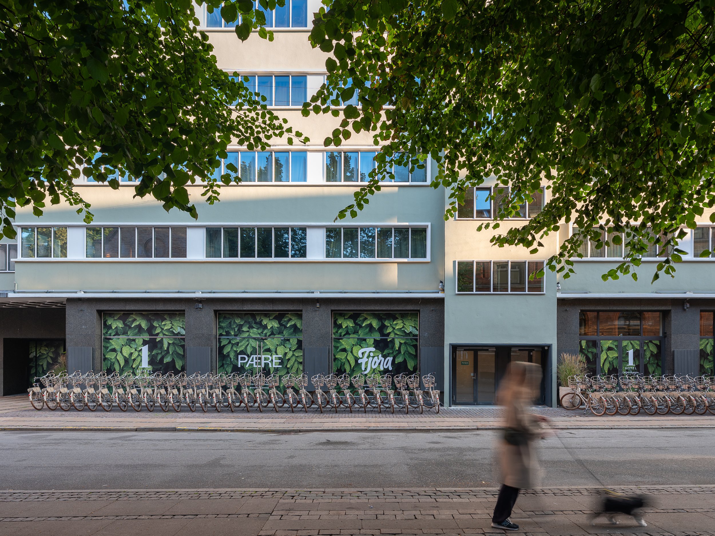 Street view of a modern building with a bike rental station in front and a person riding an electric scooter on the sidewalk; the building has large windows and green foliage at the top.