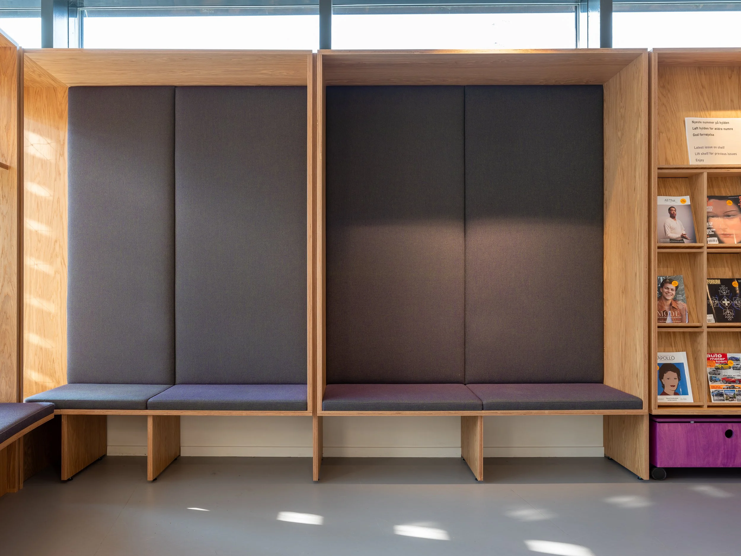 Public seating area with high-backed booths featuring dark gray cushions and wooden framing, next to a magazine rack with various magazines.
