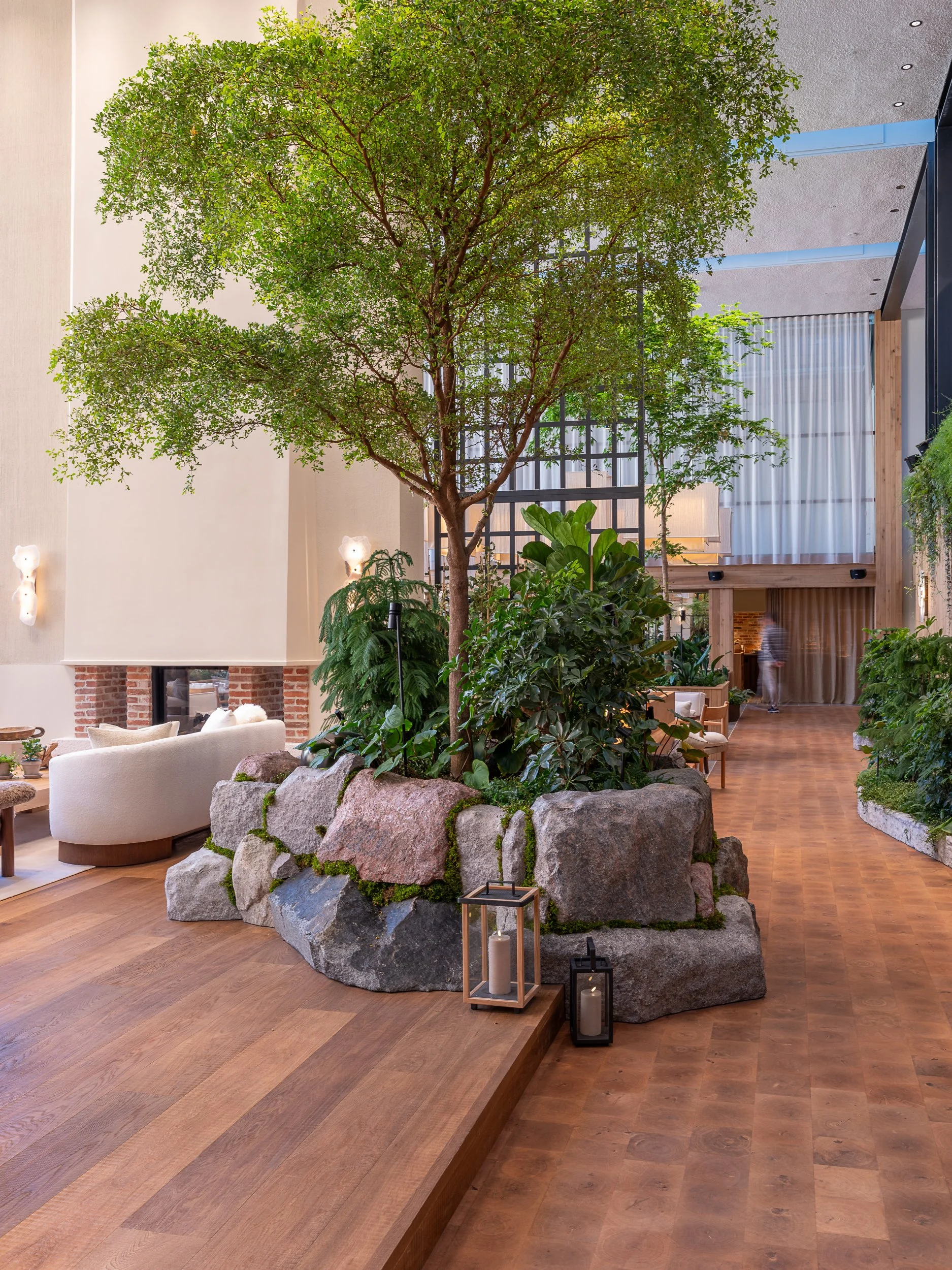 Indoor space with a large tree surrounded by rocks, plants, and candles in lanterns, with seating and a wooden floor.