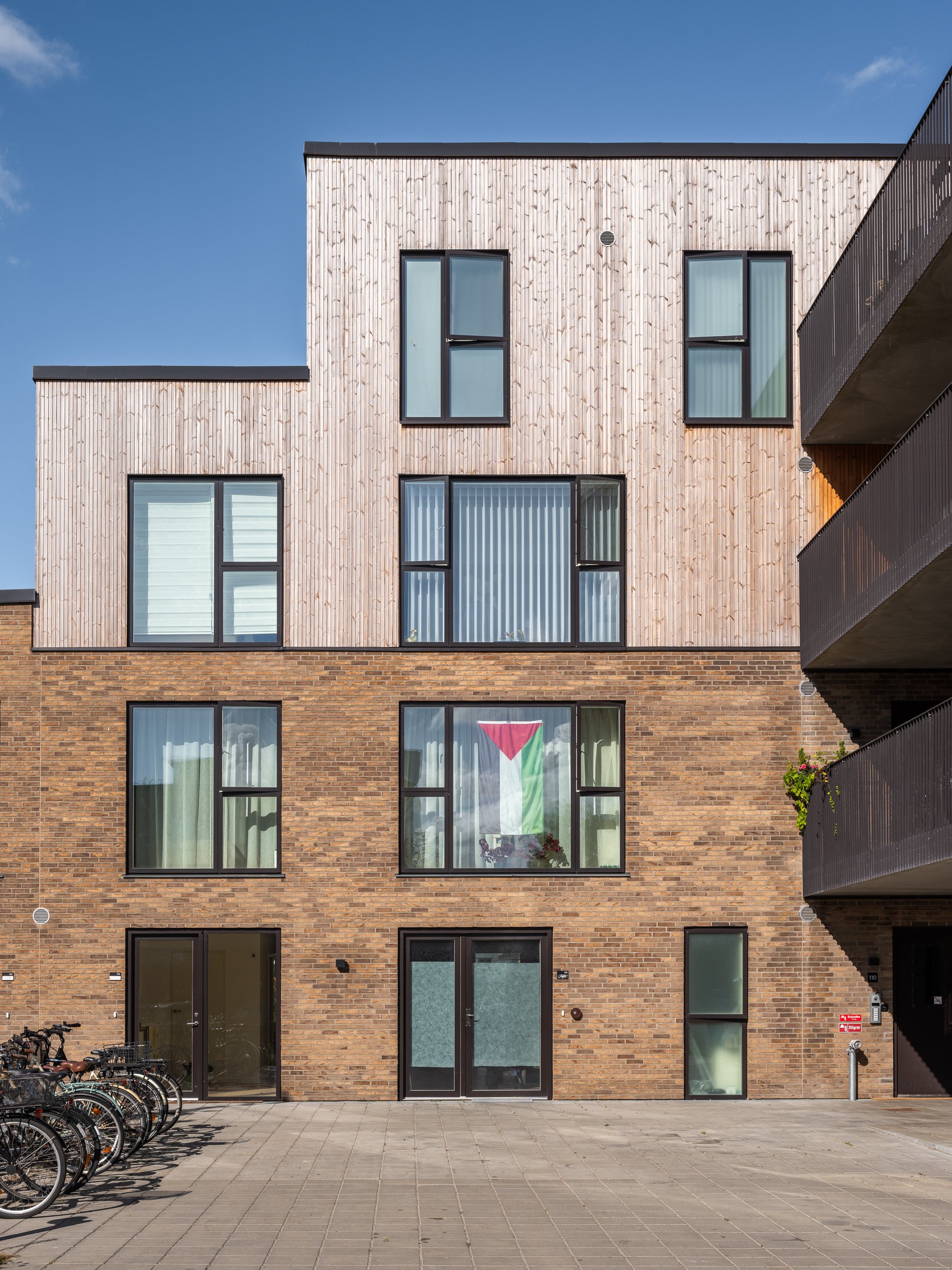 A modern four-story apartment building with a brick lower level and a light-colored wooden upper level, featuring large black-framed windows, a bicycle parking area in front, and a Palestinian flag hanging in one of the windows, under a partly cloudy