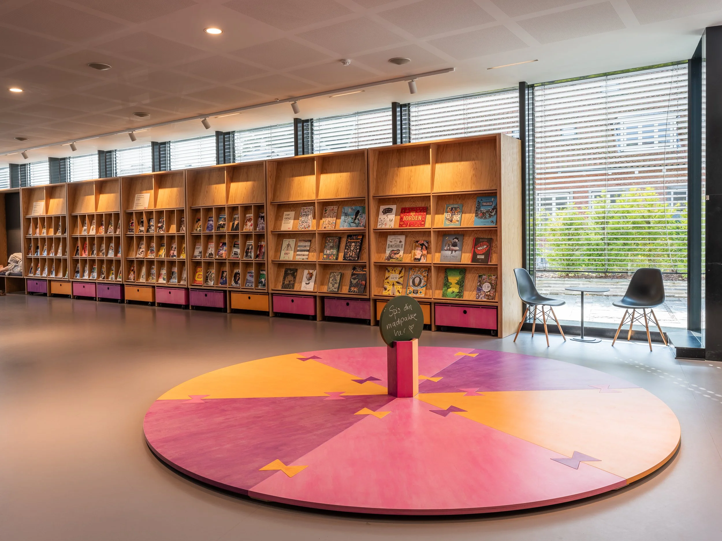 Interior of a modern library or bookstore with a colorful circular platform in the foreground, a row of wooden shelves filled with books along the wall, and two black chairs next to a small table near large windows providing natural light.