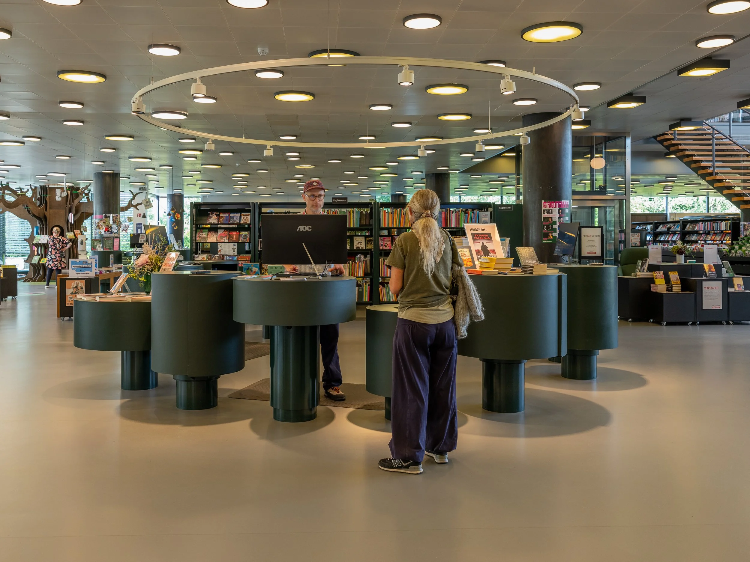 A woman standing at a library checkout counter interacting with a librarian. The library has bookshelves and a staircase in the background, with several patrons browsing.