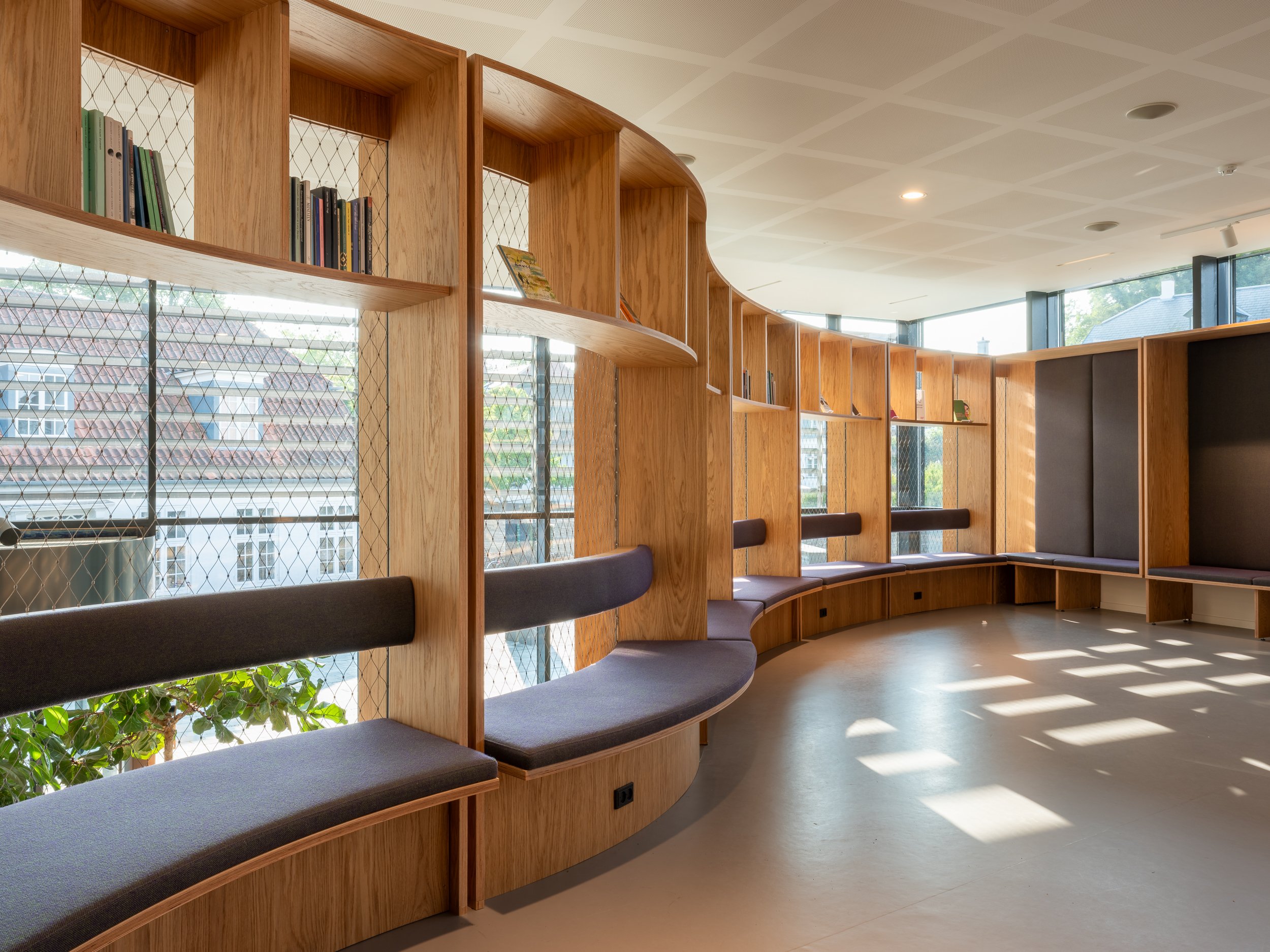 Empty modern library or reading area with curved wooden bookshelves, cushioned seating, large windows, and sunlight creating shadows on the floor.