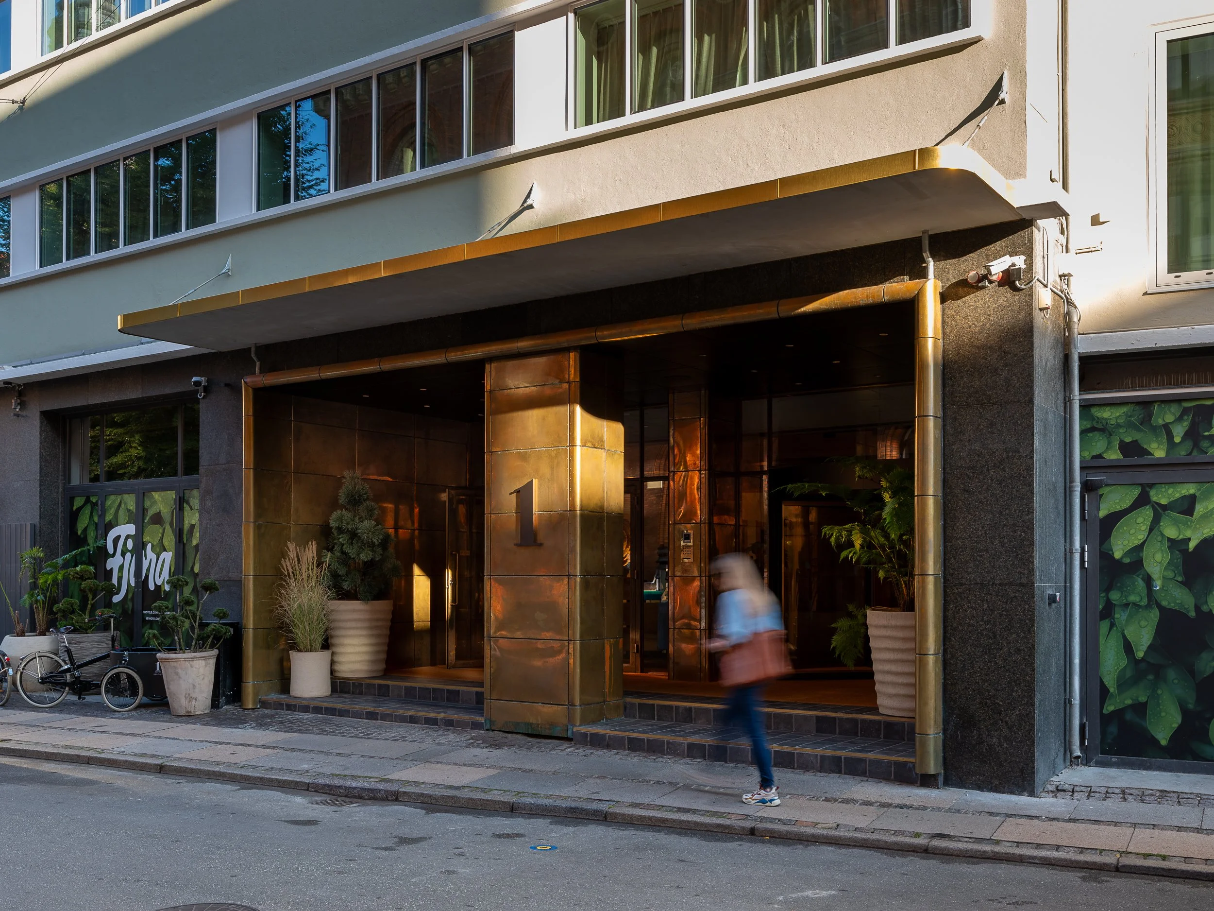 Modern building entrance with golden metallic accents, potted plants, and a person walking by.