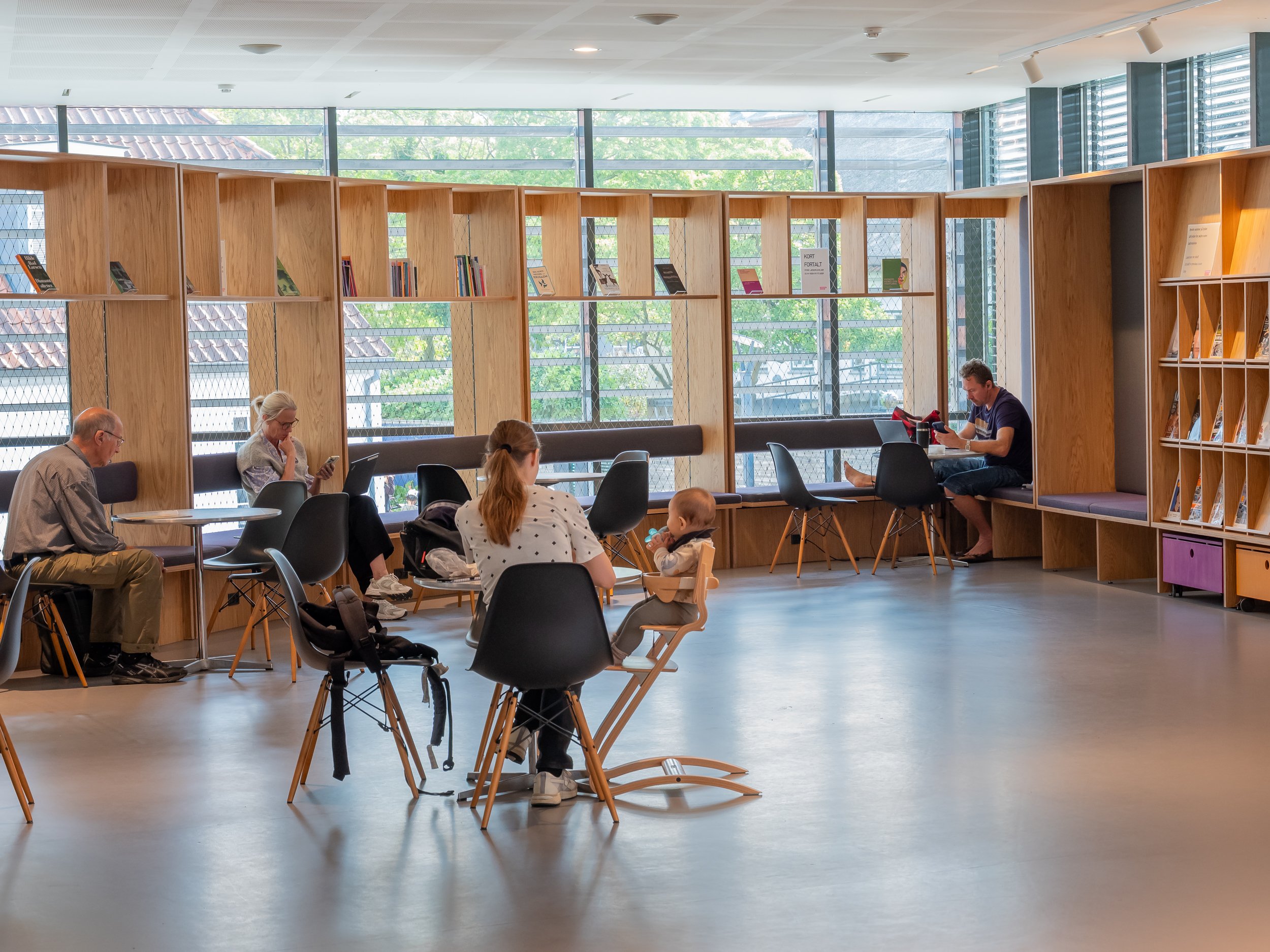 People sitting in a modern library with wooden bookshelves, some reading or using their phones, and a child sitting in a high chair.
