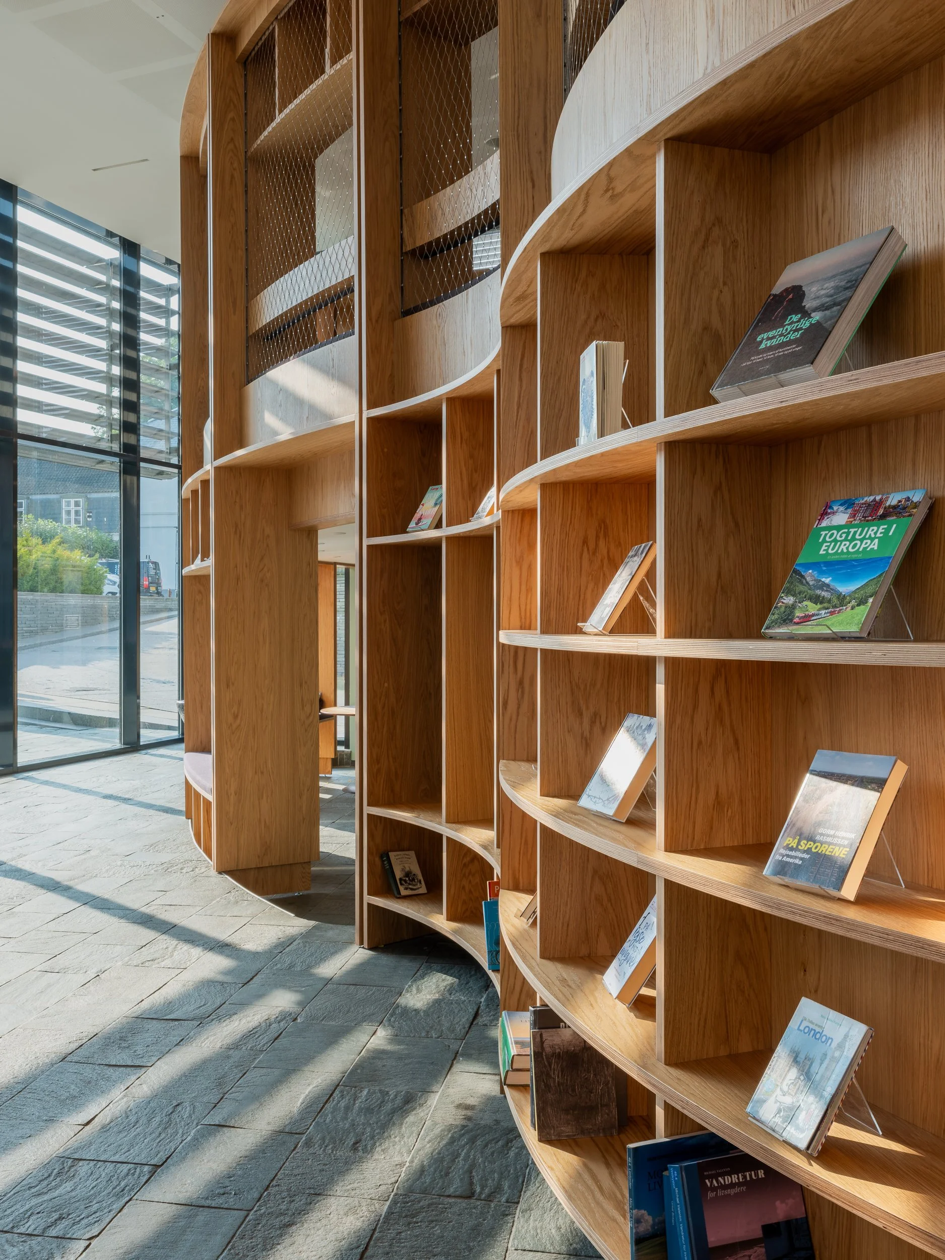 Modern wooden bookshelves with curved design in a well-lit library, holding various books, with large glass windows allowing natural sunlight.