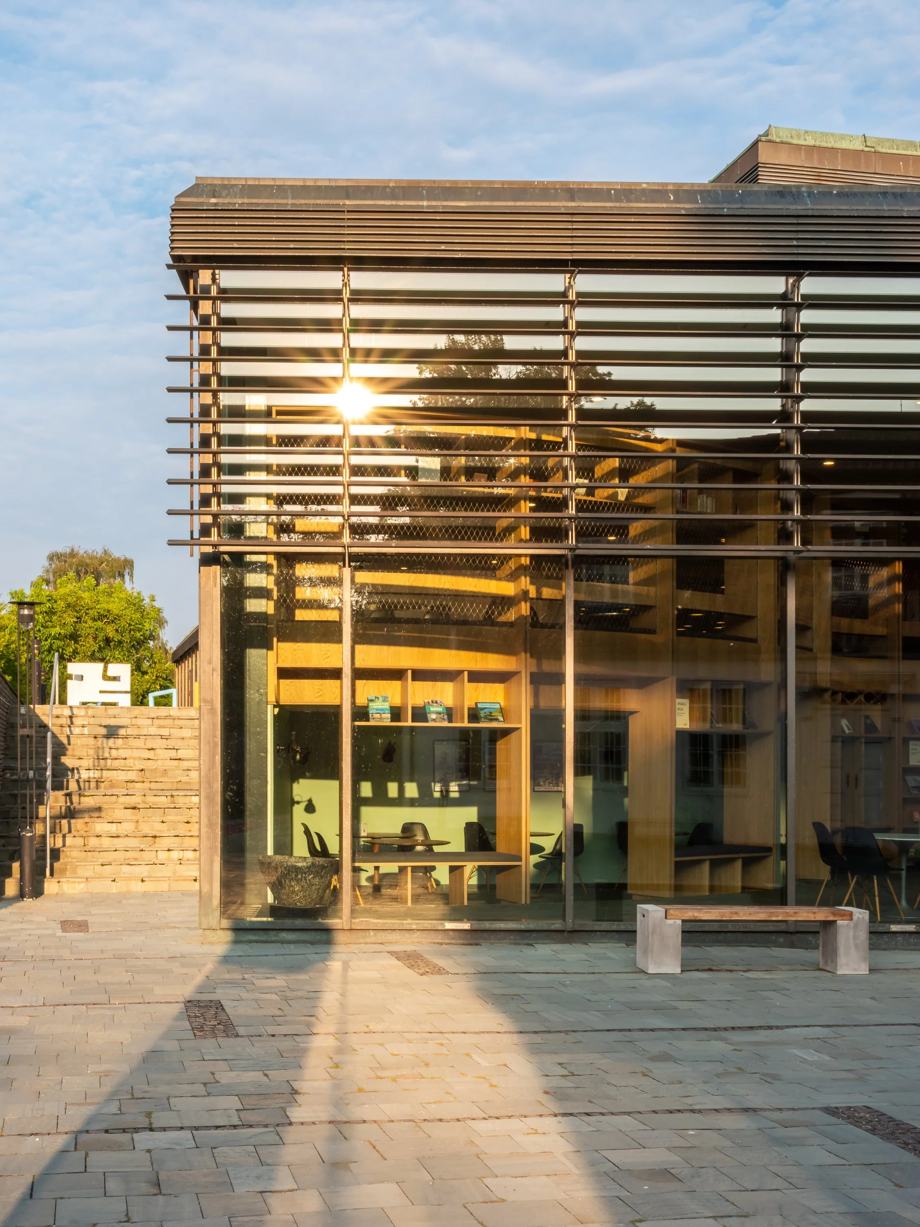 A modern building with glass walls and horizontal slats, sunlight reflecting off the glass, with outdoor stairs, a concrete bench, and a clear sky.