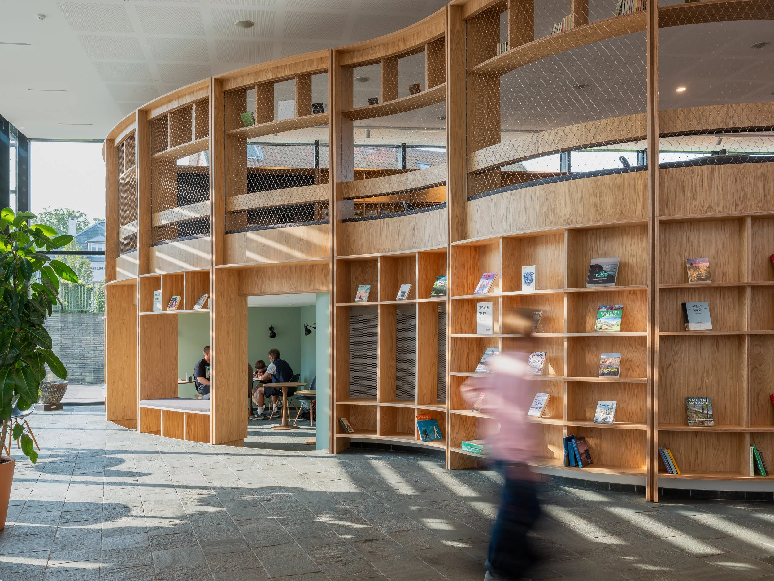 Modern interior with curved wooden bookshelf filled with books, a person walking by, and a seating area with people working at a table.