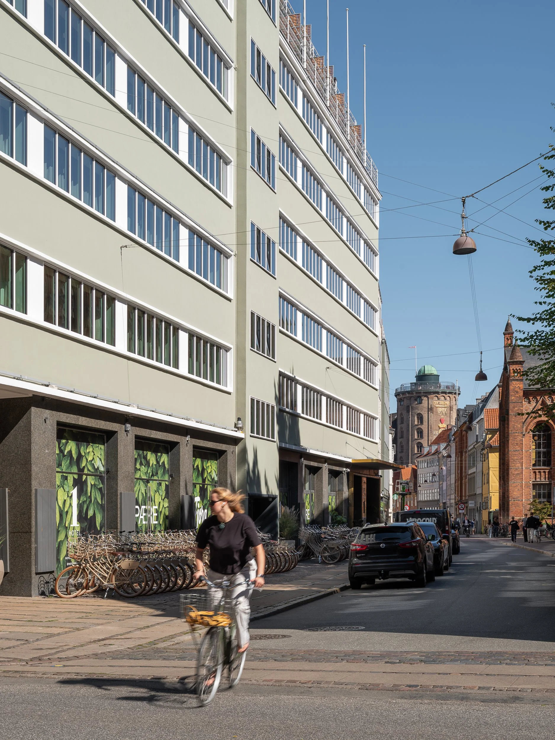 A city street scene with a woman riding a bicycle past a modern light-colored building with large windows. There are parked cars and bicycles along the curb, with historic brick buildings and a tower in the background under a blue sky.