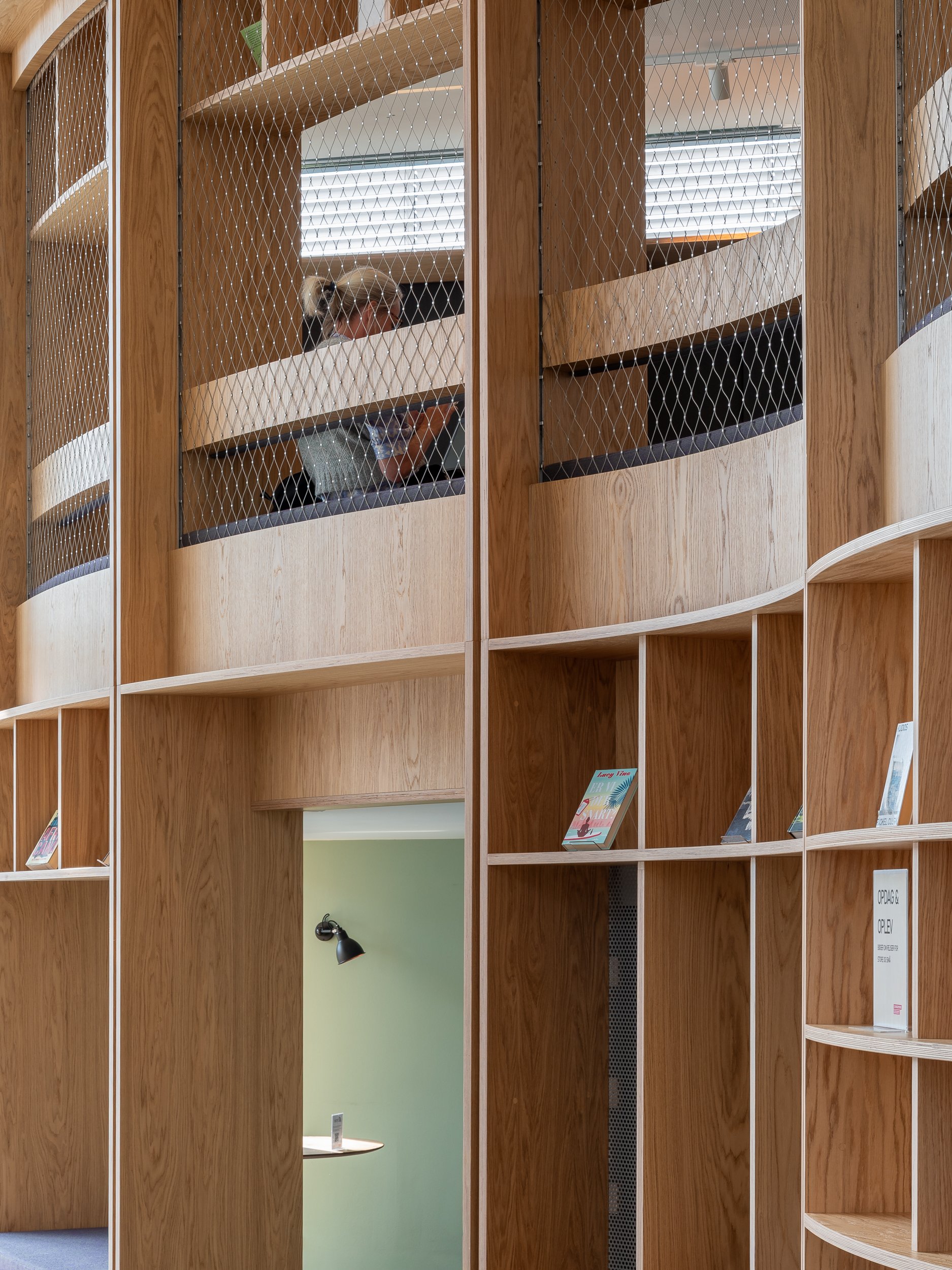 A woman with blonde hair tied in a bun, seen through a window with wire mesh, sitting in a room with green walls, a black wall lamp, reading or working at a round white table.