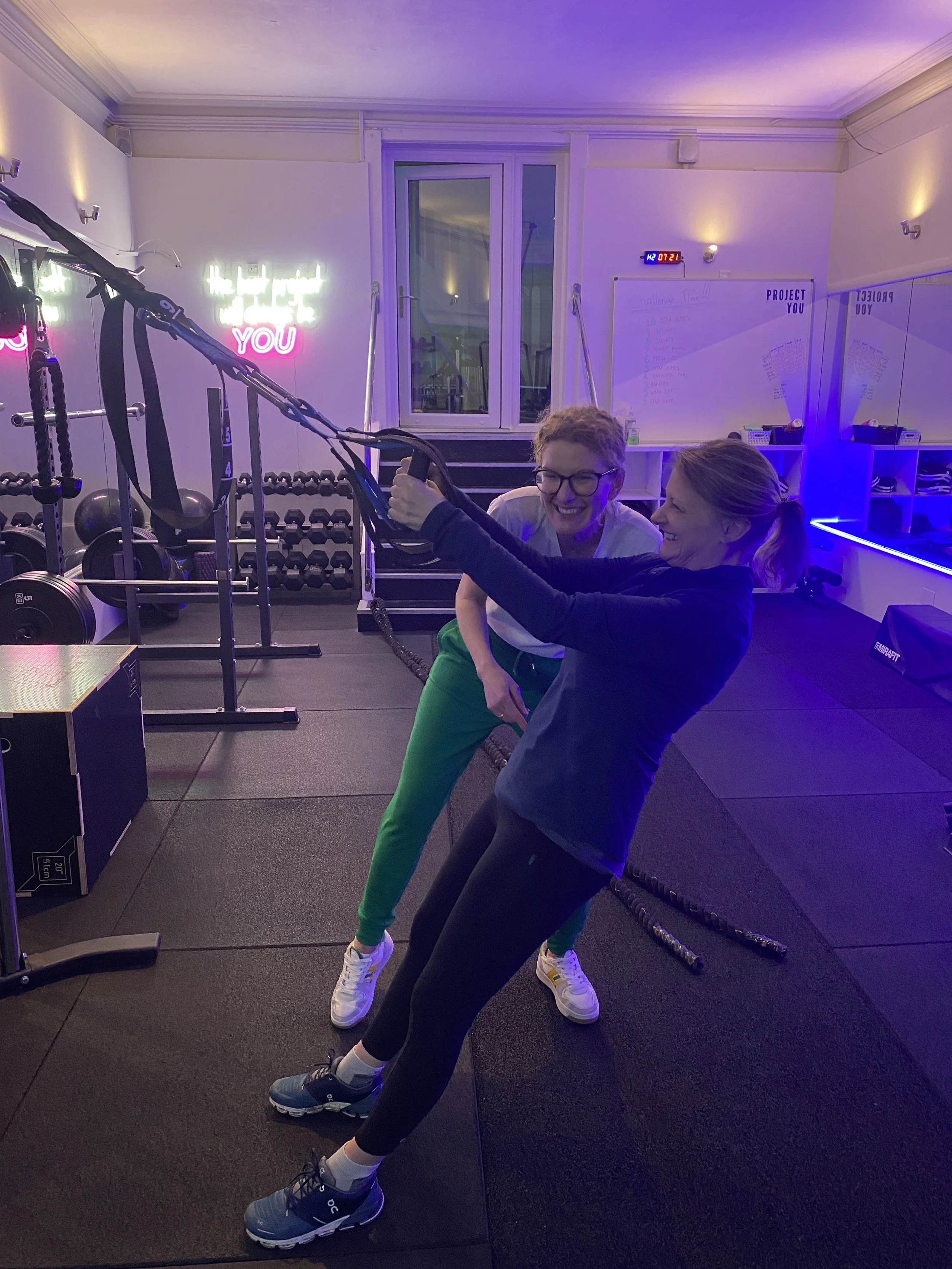 Two women working out in a gym, one holding a suspension trainer and the other assisting, both smiling. The gym has black mats, weight racks, and neon signs on the wall.