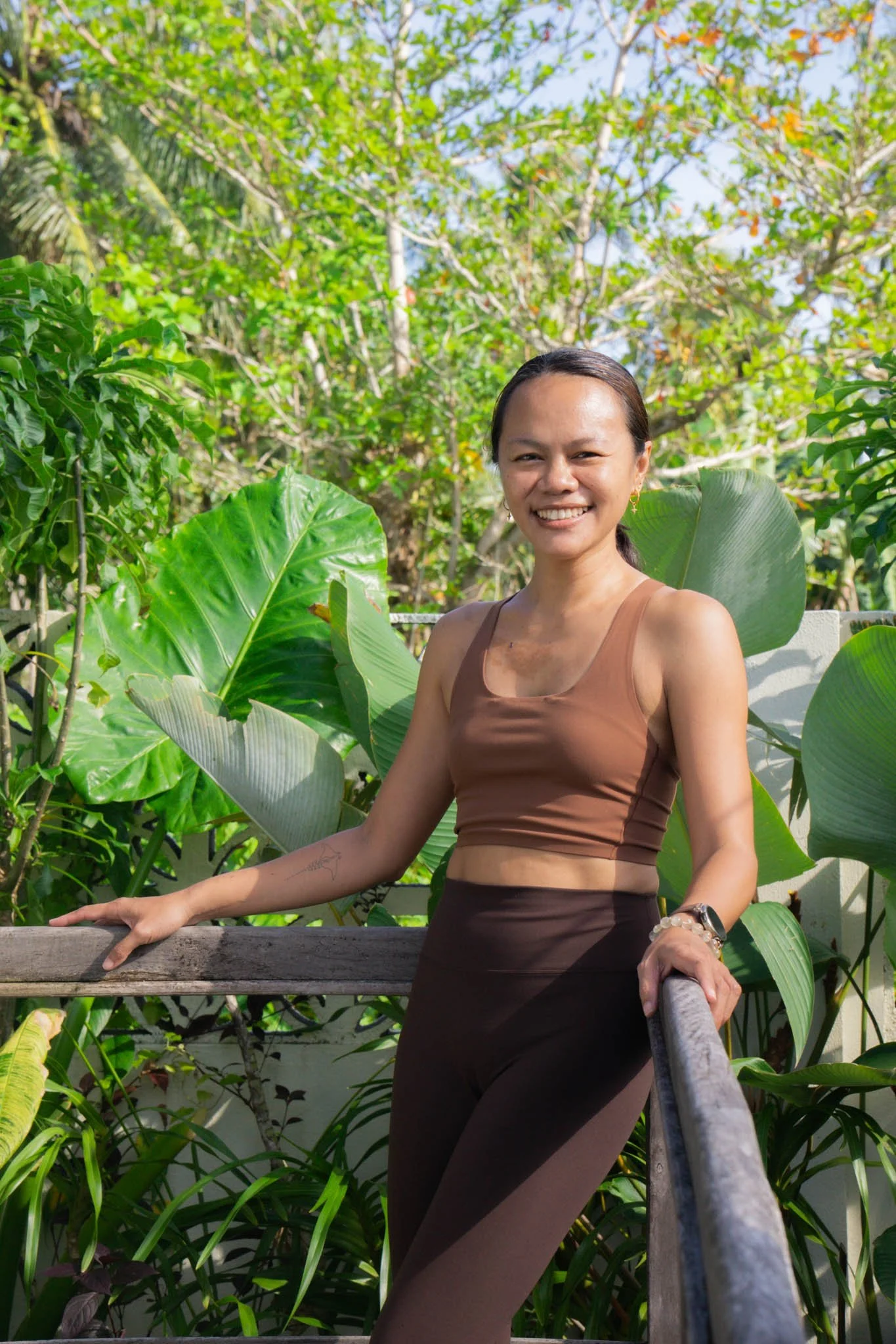 A woman with dark hair pulled back, smiling, wearing a brown sports bra and matching high-waisted leggings, standing on a wooden railing outdoors surrounded by green tropical plants and trees.