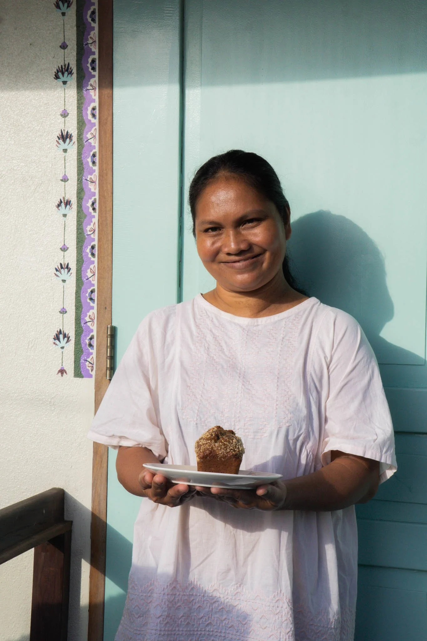 Woman holding a plate with a muffin, standing in front of a blue door and a decorative wall hanging.