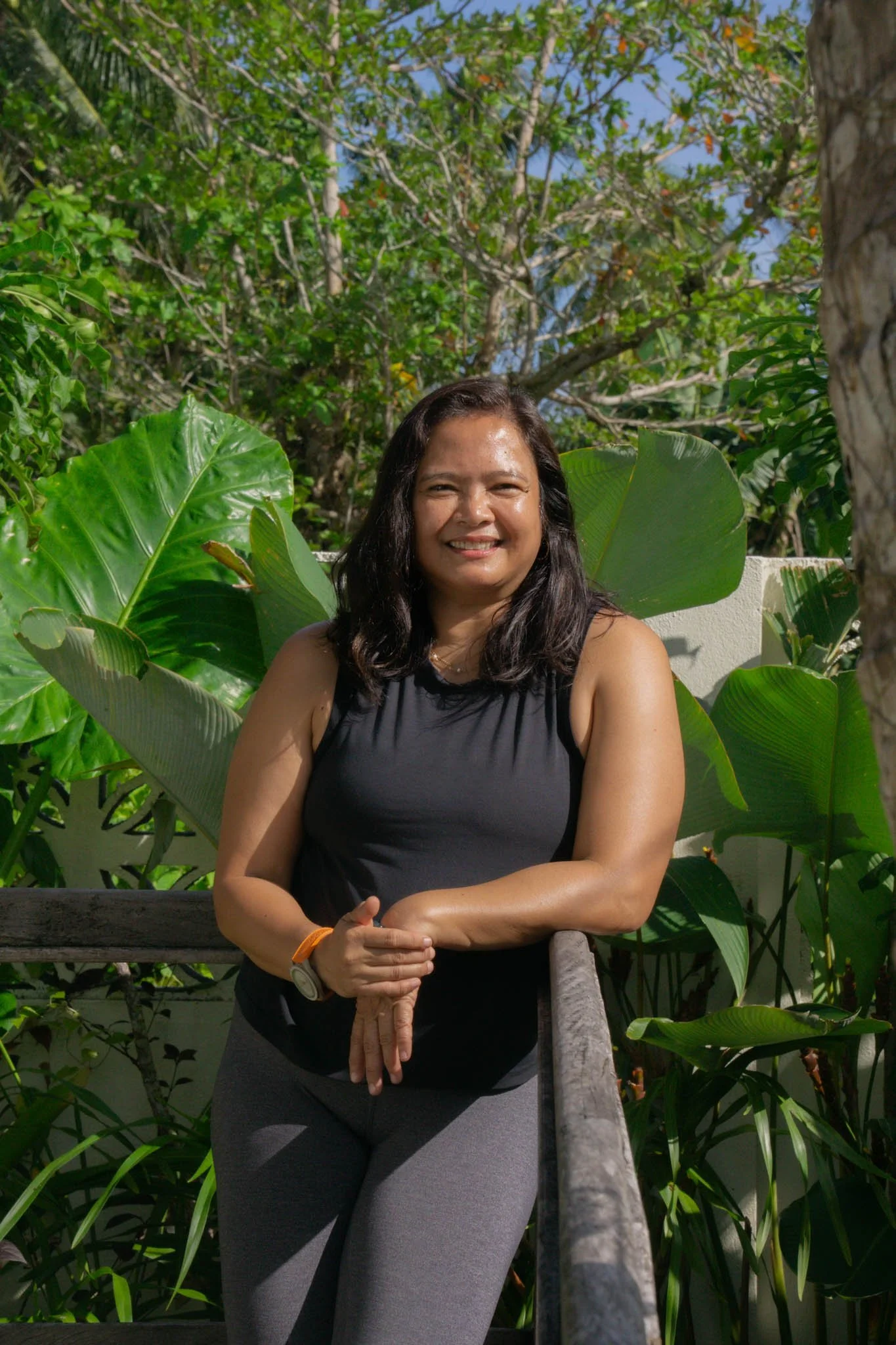 A woman with dark hair smiling outdoors, standing near large green tropical leaves and trees.
