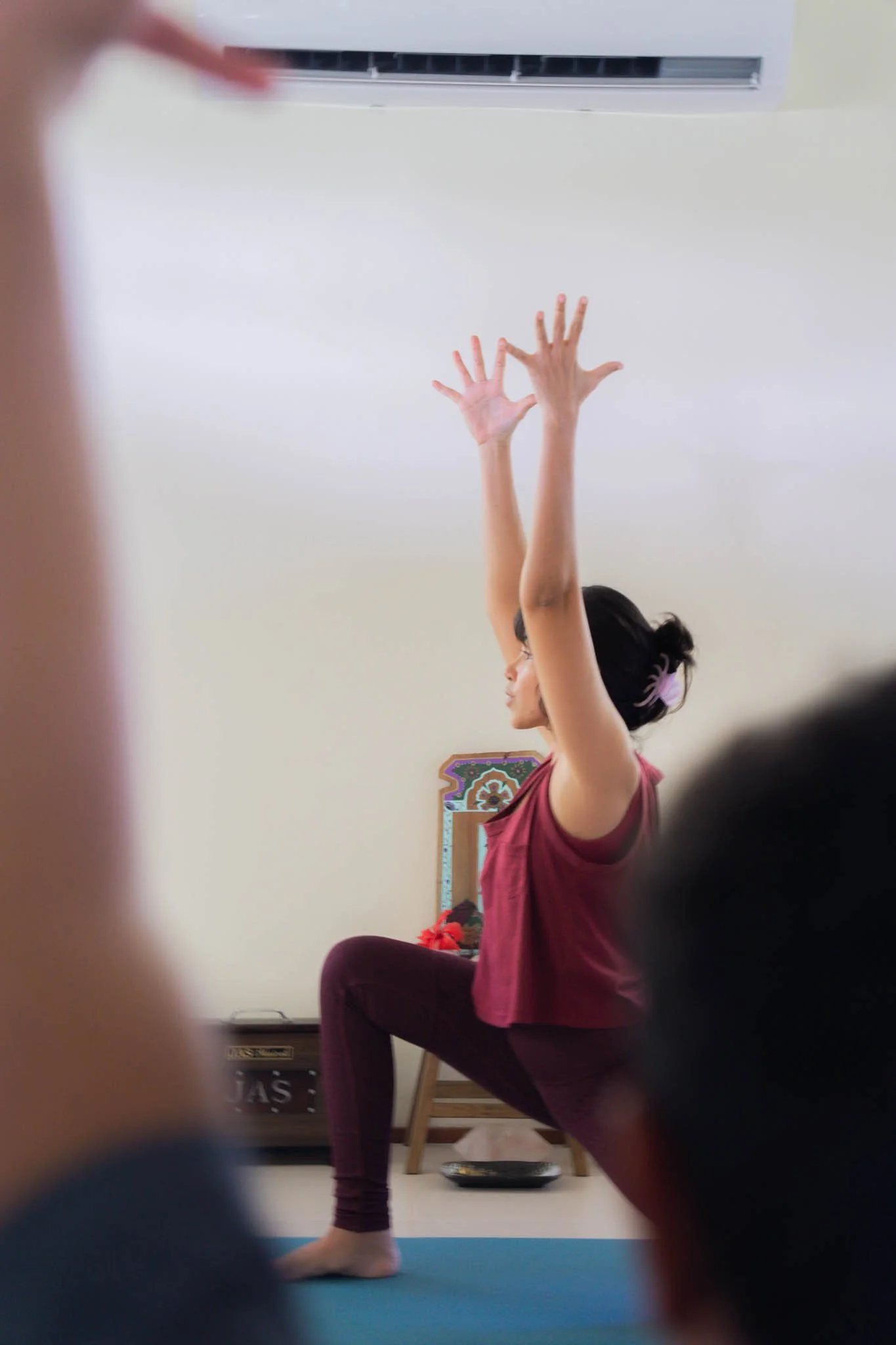 A woman practicing yoga or stretching indoors, seated on a mat with arms raised overhead, surrounded by other people.