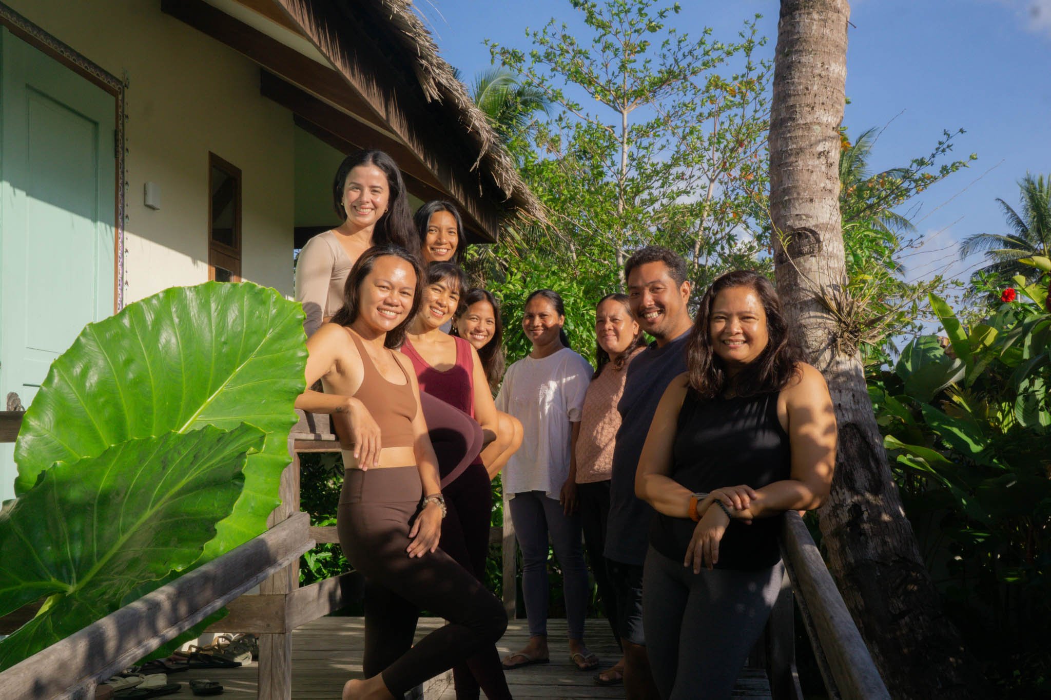 A group of ten diverse people smiling outdoors on a sunny day, standing on a wooden porch surrounded by lush green tropical plants and trees, next to a light-colored house with a thatched roof.