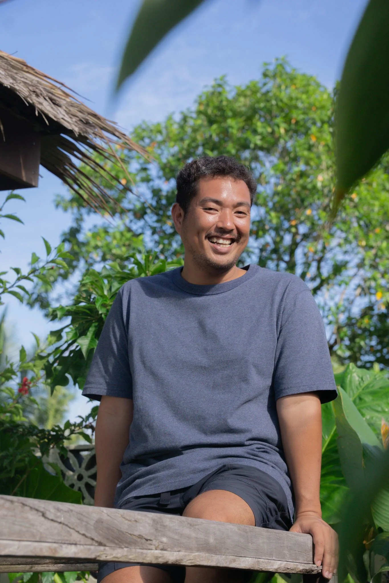 A man smiling outdoors, sitting on a wooden bench surrounded by lush green plants and trees, with a thatched roof structure and clear blue sky in the background.