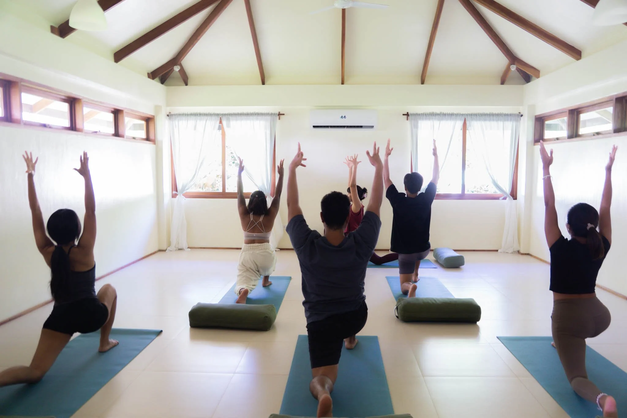 A group of six people practicing yoga in a bright room with large windows, some performing a lunge pose with arms raised.