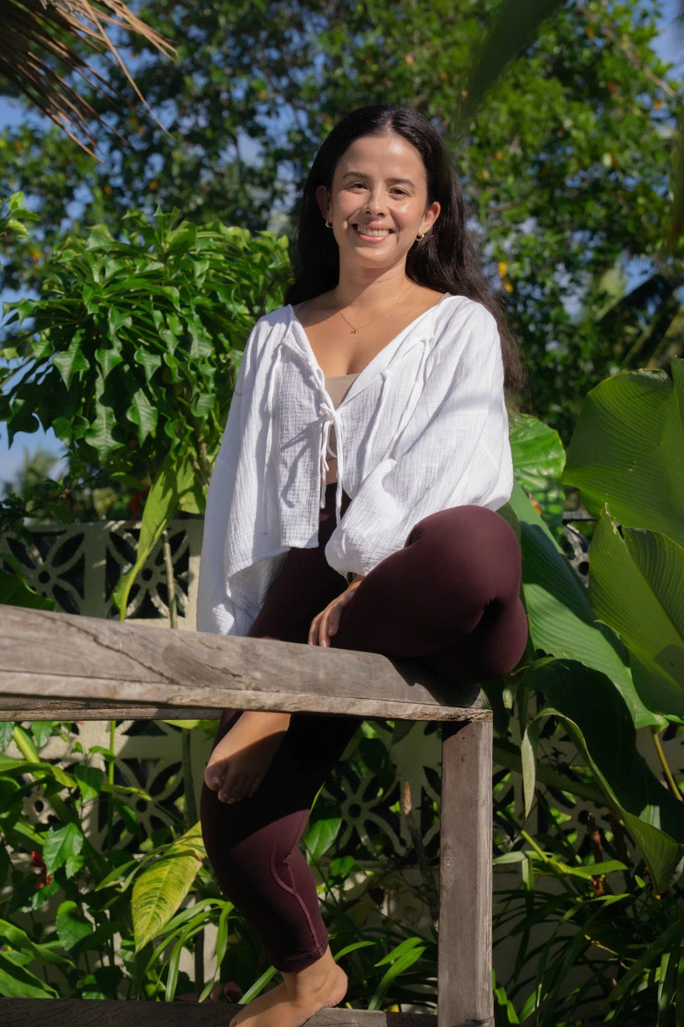 A woman smiling outdoors sitting on a wooden surface, surrounded by green tropical plants and trees, wearing a white blouse and maroon leggings.
