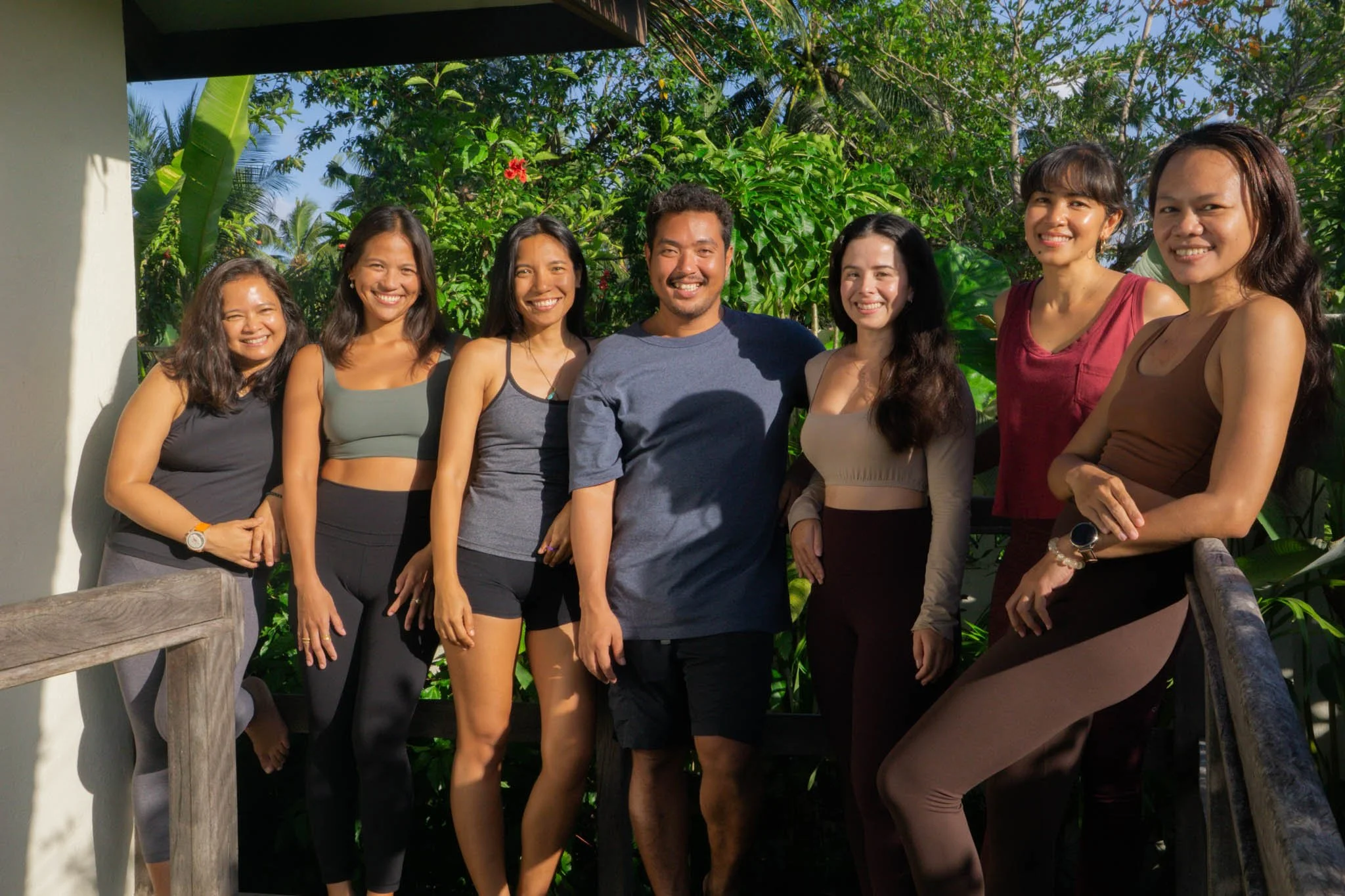 Group of eight smiling people standing outdoors with lush green trees in the background.