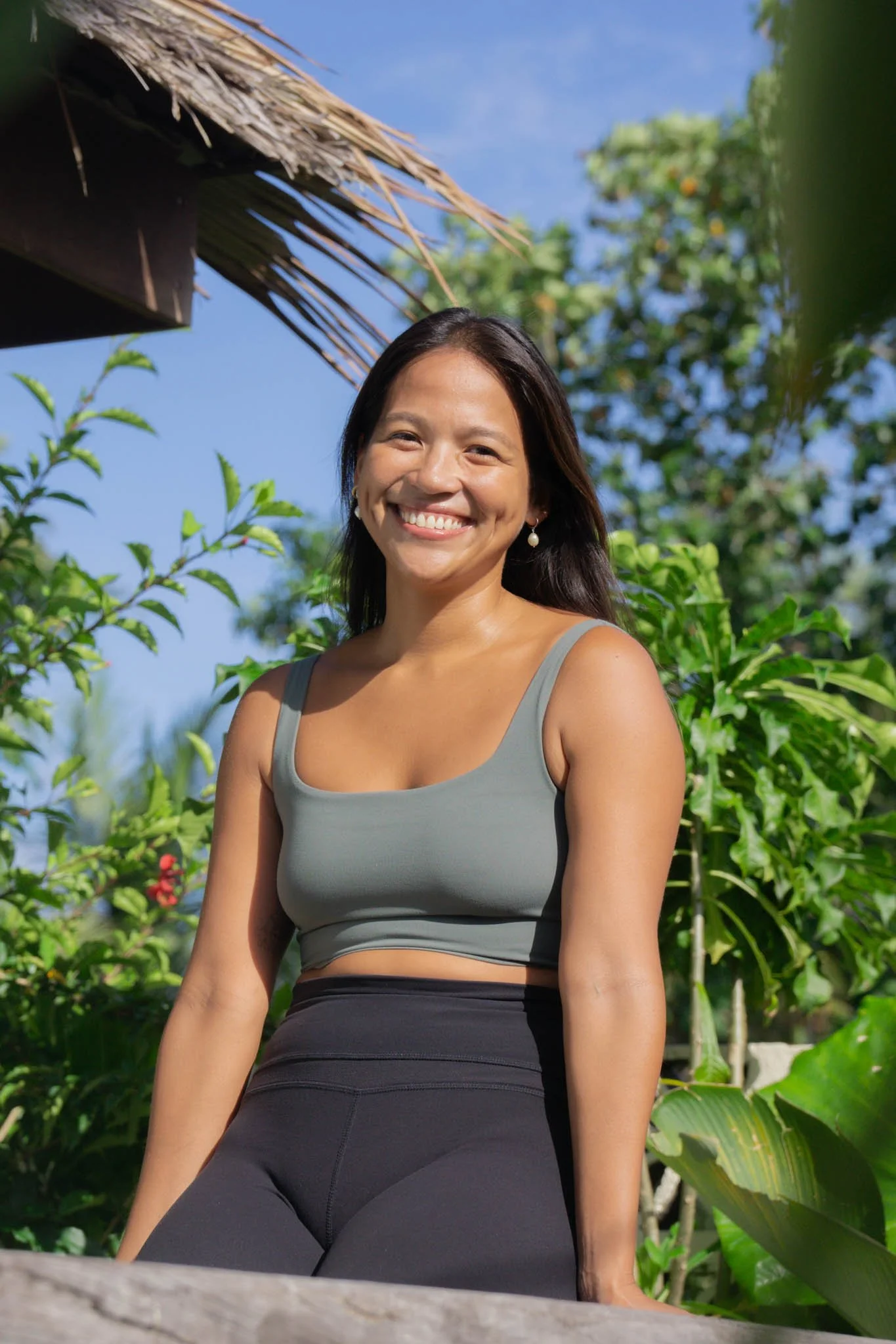 A woman smiling outdoors in a sunny environment with green foliage and a thatched roof in the background.