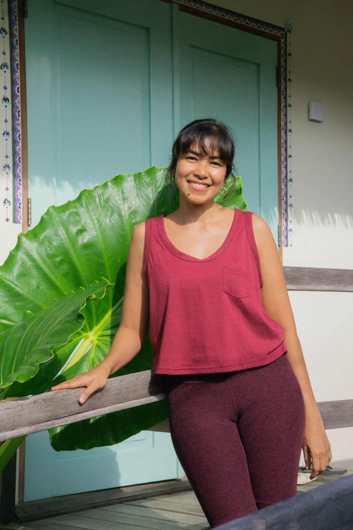 A woman in a red sleeveless top and maroon leggings is smiling and leaning on a wooden railing next to large green leaves outside a building with a teal door.