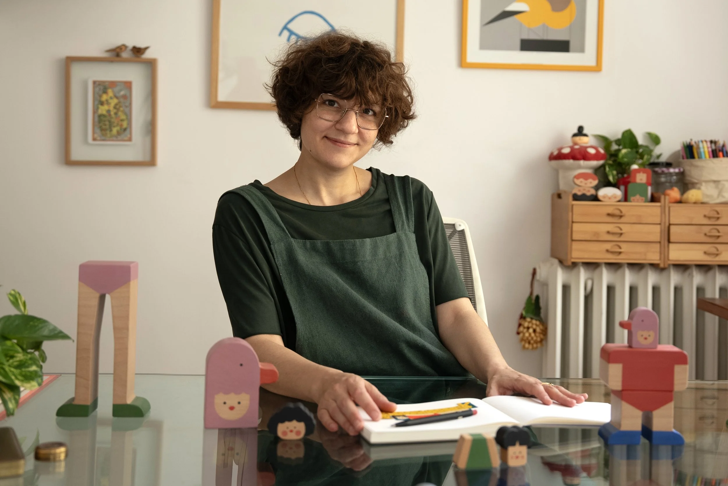 A woman with short curly hair and glasses sitting at a glass table, surrounded by colorful wooden toys, with framed artwork on the wall behind her.