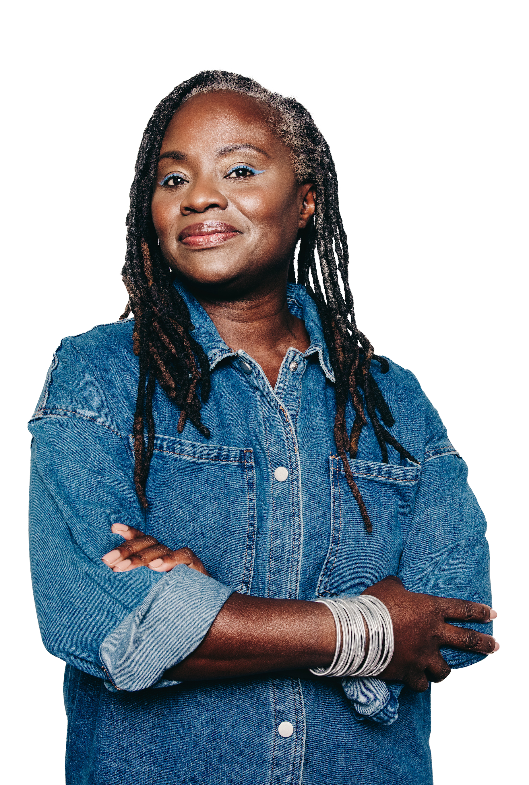 A woman with long dreadlocks, wearing a denim shirt and multiple silver bangles on her wrist, stands confidently with her arms crossed.