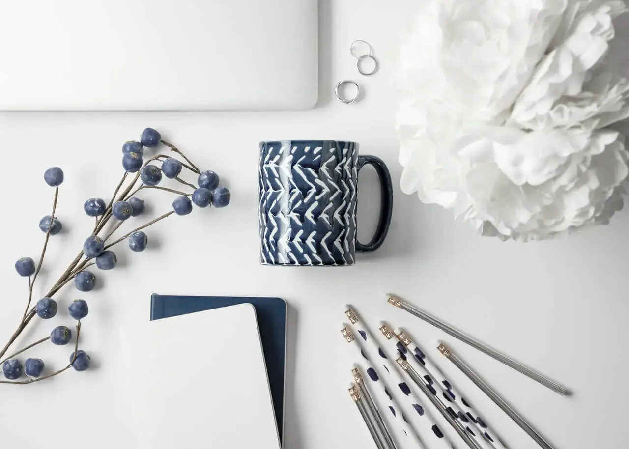 Flat lay of office supplies and decor on a white surface, including a patterned mug, a small branch with blue berries, a white flower, a laptop, notebooks, pens, and a noose.