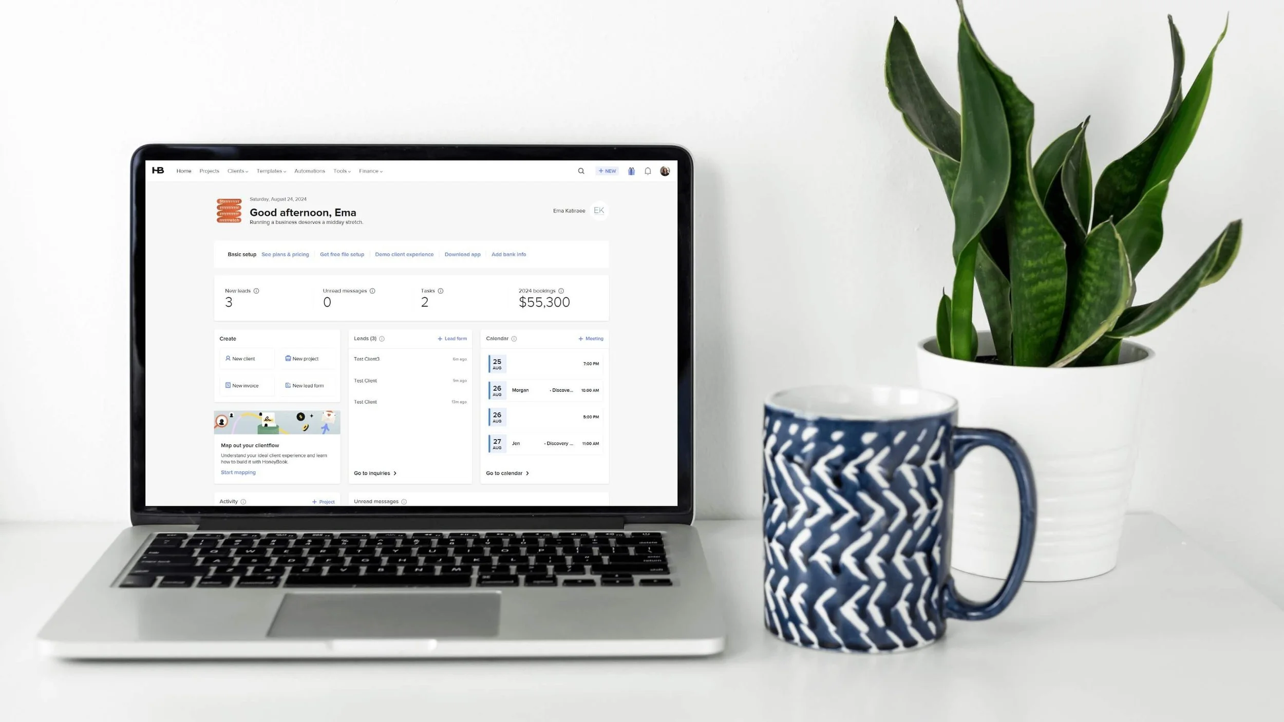 A laptop displaying HoneyBook dashboard sits on a white desk next to a potted snake plant and a blue and white patterned coffee mug.