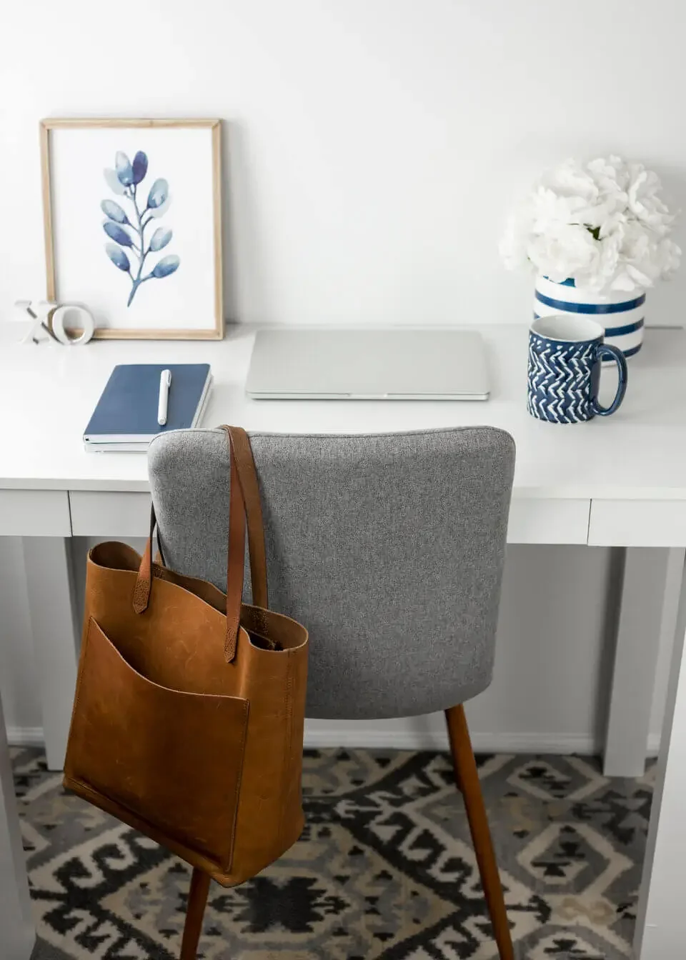 Modern white desk with laptop, navy notebook and pen, framed botanical art, flower vase with white flowers, patterned mug, gray chair, brown leather tote bag hanging on the chair, and a patterned rug.