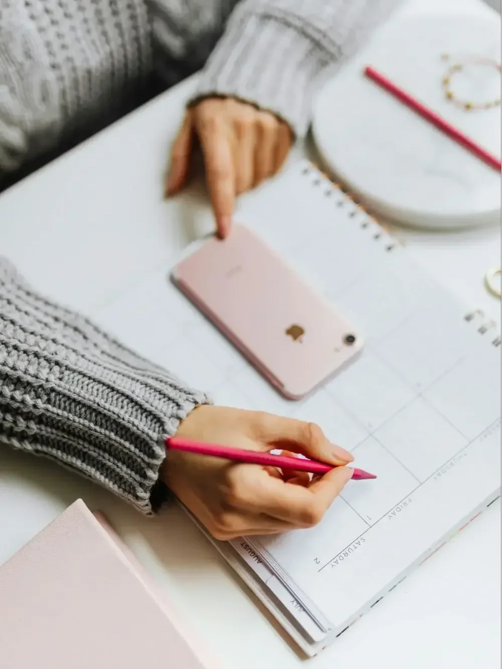 A person writing in a planner with a pink pen, wearing a gray sweater. On the table, there is a pink, a white notebook, and a bracelet