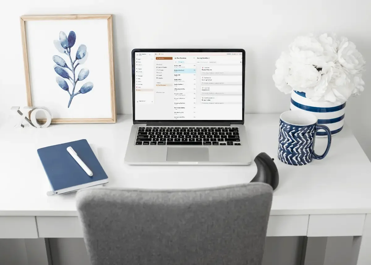 A clean white desk with a laptop displaying an email inbox, a blue notebook with a white pen, a striped blue and white mug, a large vase of white flowers, and a framed watercolor of blue leaves, in a bright, minimalist workspace.