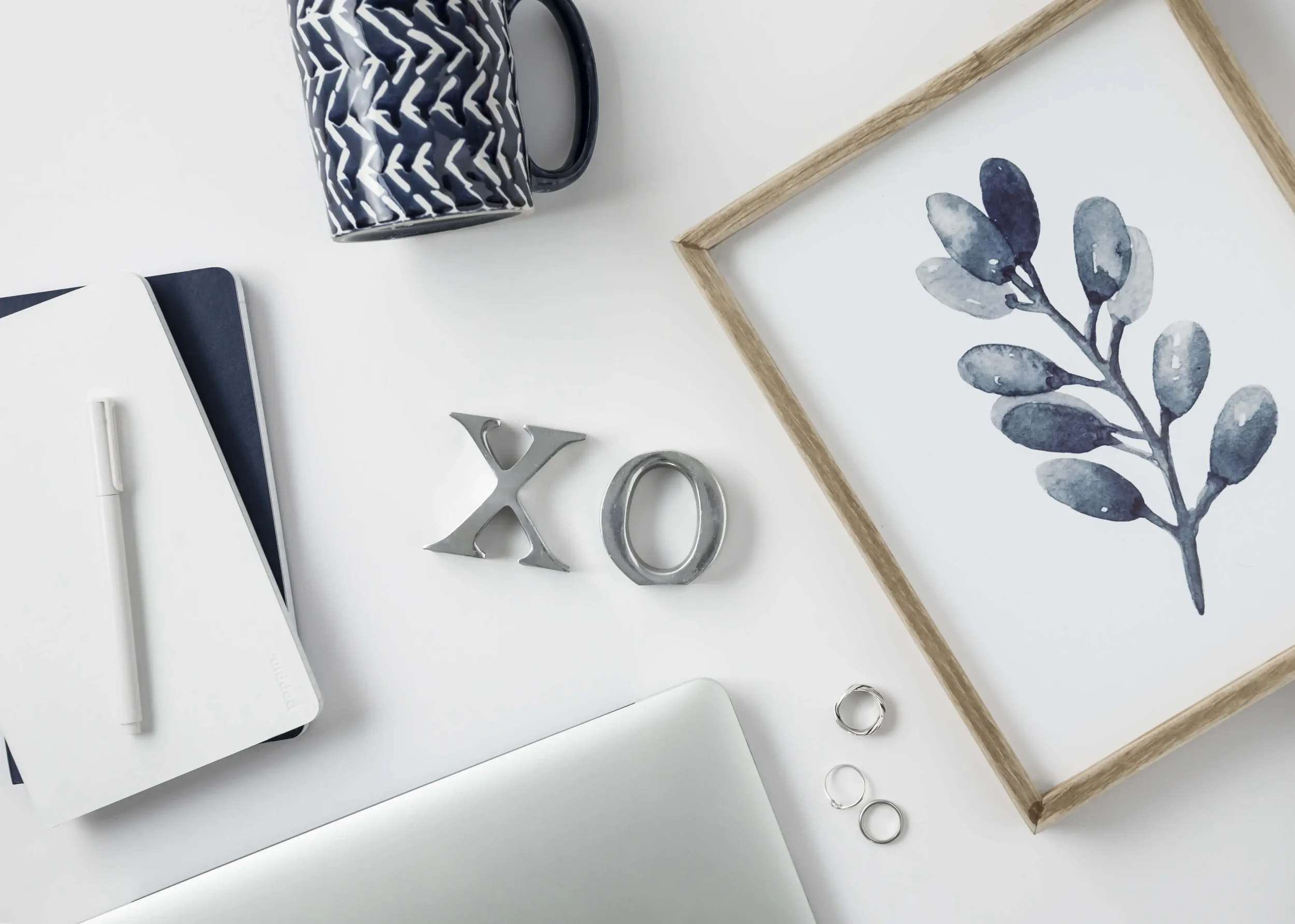 Flat lay of a white workspace with a black and white zigzag mug, a closed white notebook, a silver pen, metallic 'X' and 'O' character figurines, and a watercolor floral illustration in a wooden frame.