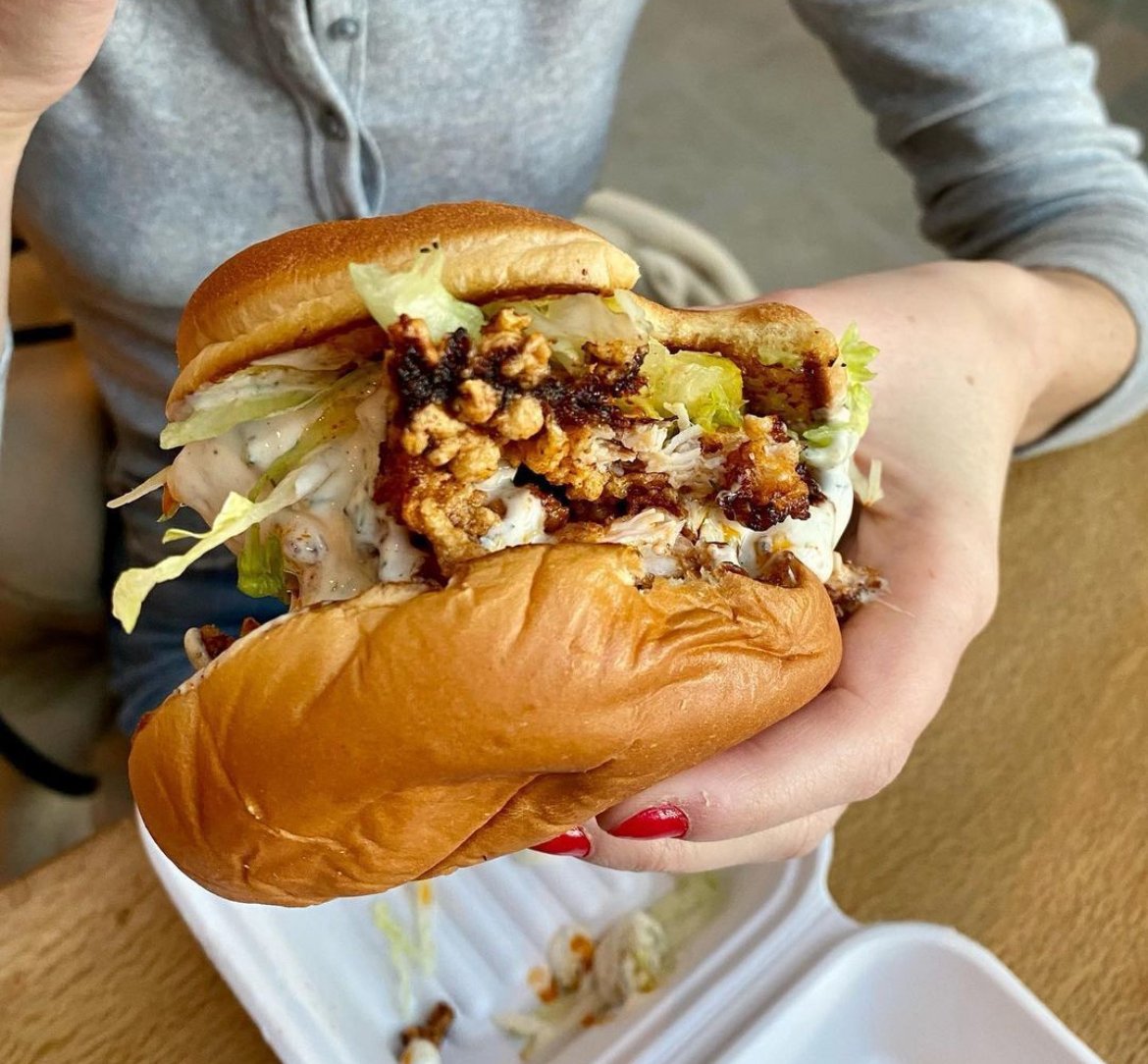 Person holding a large sandwich with fried chicken, lettuce, mayo, and pickles on a bun.
