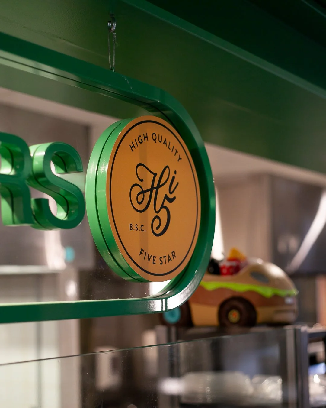 Close-up of a round yellow sign with a green border that reads 'High Quality Five Star' and 'Hi B.S.C.' with a decorative 'Hi' in the center, hanging from a green shelf, with a blurred background of a toy car.