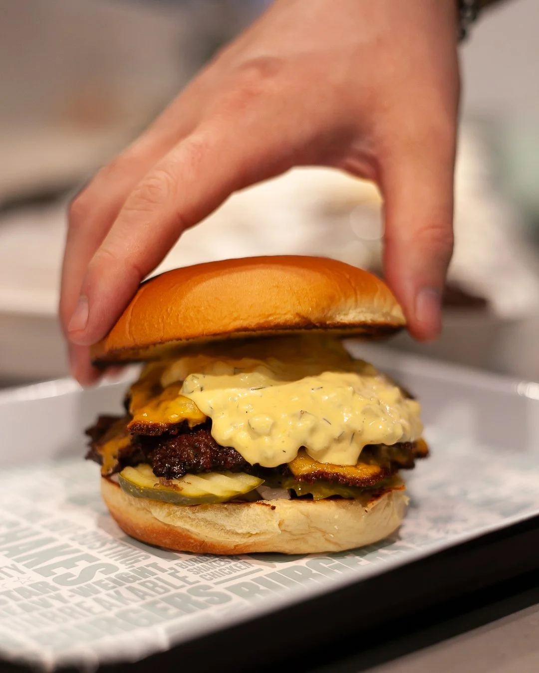 Close-up of a cheeseburger with pickles, bacon, and sauce, being placed on a paper-lined tray by a person's hand.