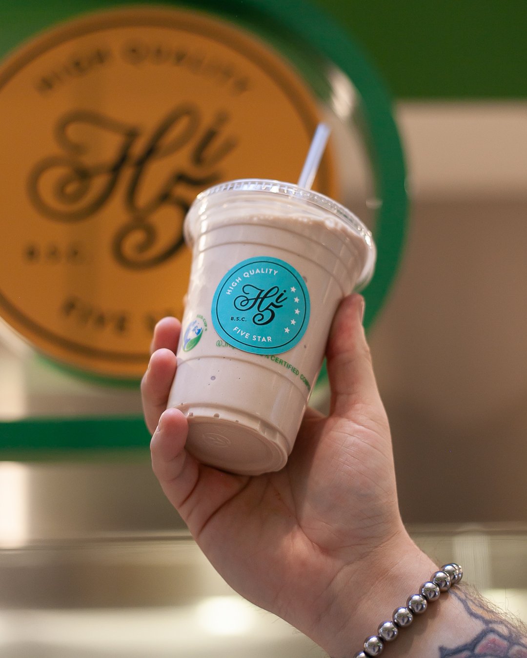 A hand holding a plastic cup of a light-colored beverage with a straw, in front of a blurred sign with the word 'Hi' and a green background.
