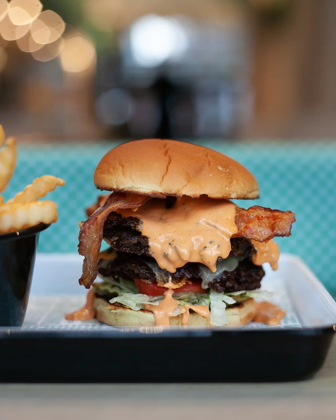 Close-up of a cheeseburger with bacon, lettuce, tomato, melted cheese, and sauce on a bun, served with a side of French fries in a black cup.