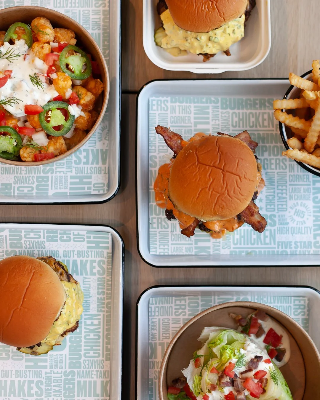 Two cheeseburgers with bacon on buns, a bowl of fries, a bowl of fried cauliflower with jalapeño slices, a salad with lettuce, tomatoes, bacon, and dressing, and a cup of potato salad with chopped vegetables.