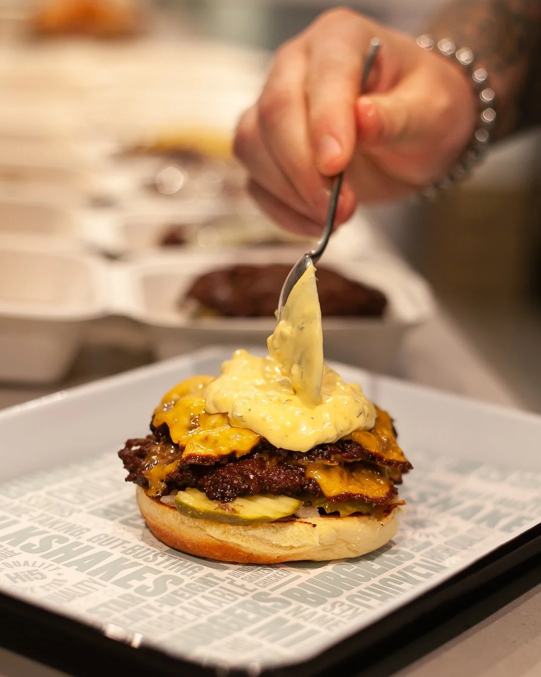 A person is spooning a creamy yellow sauce onto a burger with meat, cheese, and pickles on a bun, on a tray with printed paper.