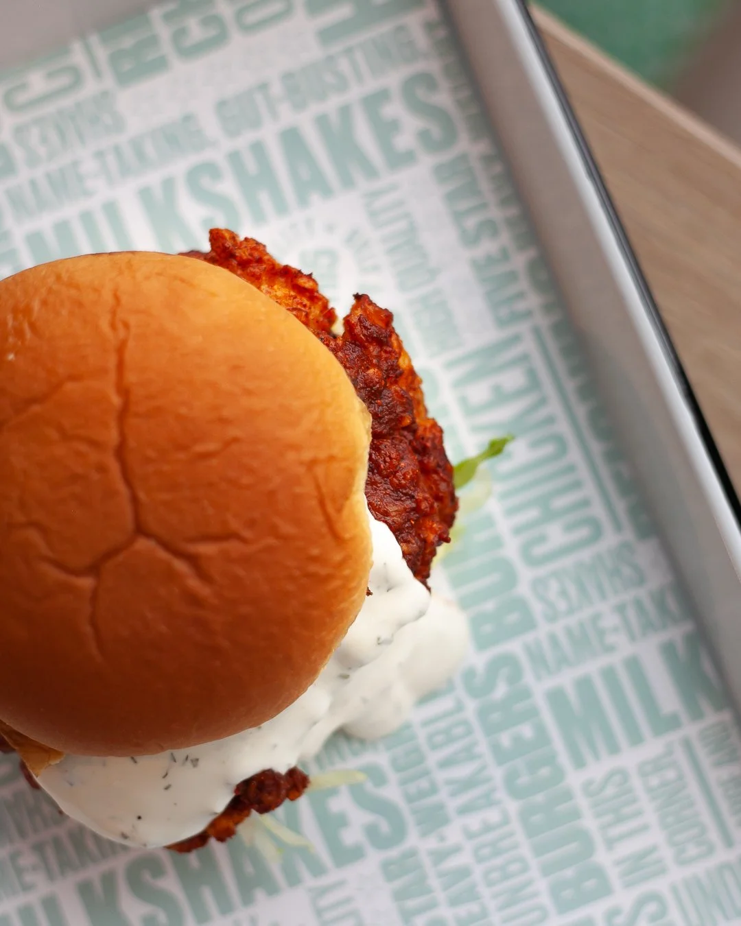 Close-up of a sandwich with a toasted bun and fried chicken fillet, topped with mayonnaise on a tray lined with branded paper.