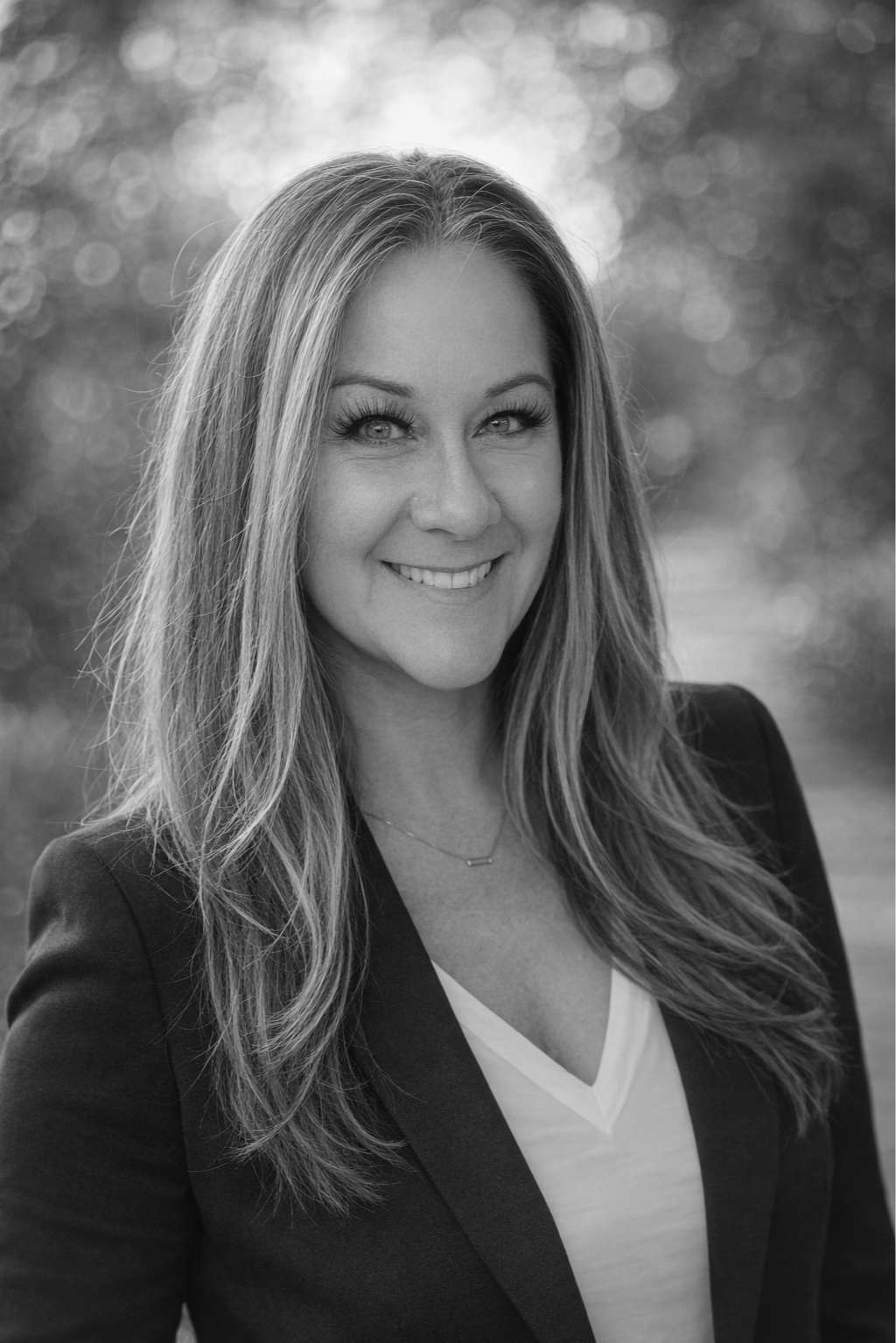 A black-and-white portrait photograph of a woman with long hair, smiling, outdoors with blurred background.