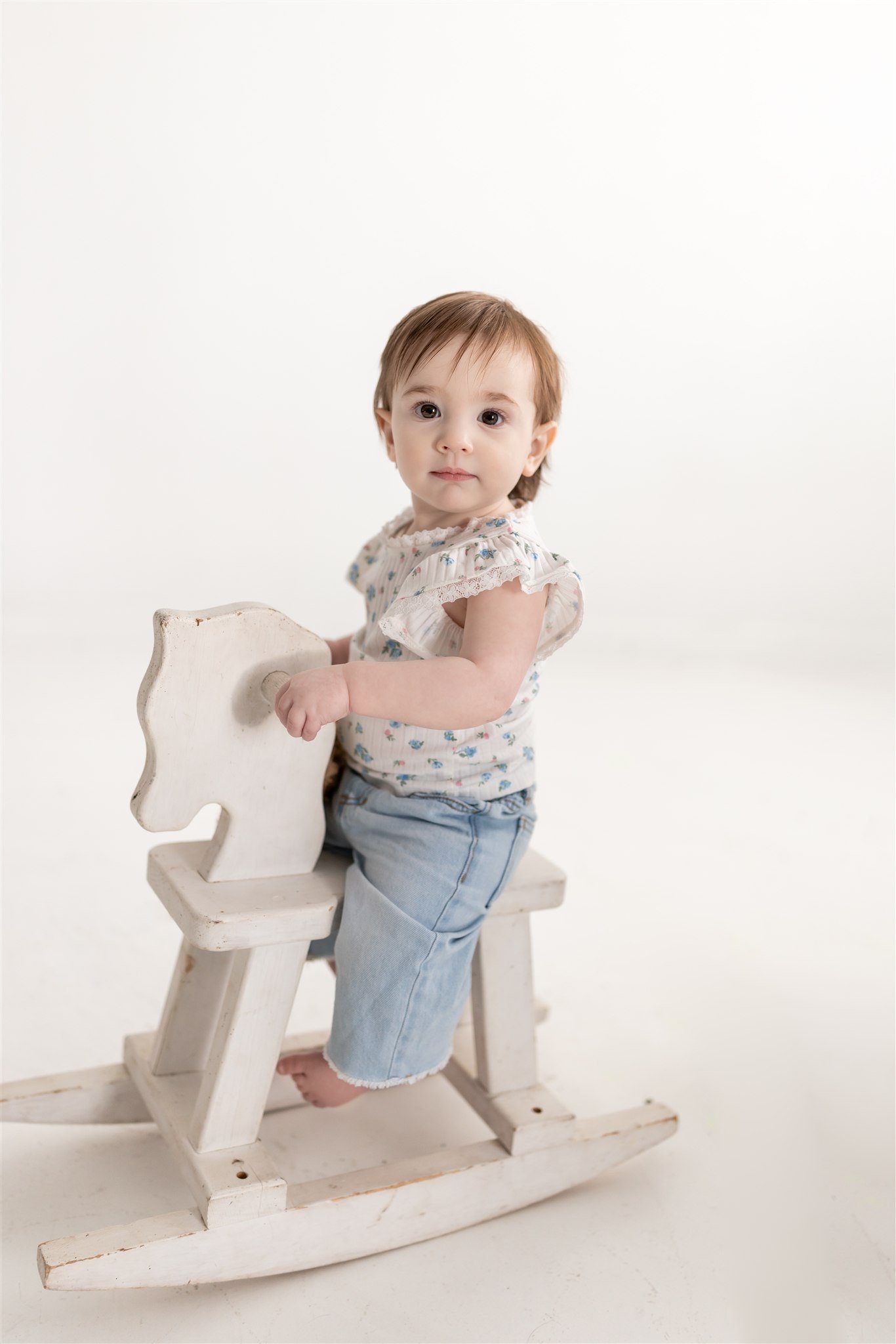 A young child with brown hair sitting on a white wooden rocking horse, wearing a floral top and light blue denim pants, against a plain white background.