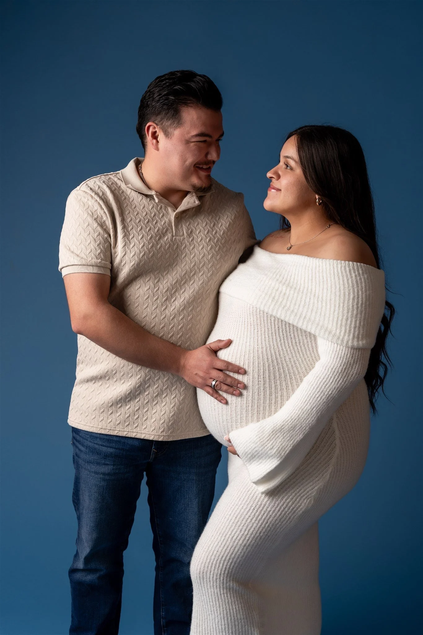 A pregnant woman with dark hair in a white sweater and her partner, a man with dark hair in a beige shirt, standing close and smiling at each other against a blue background.
