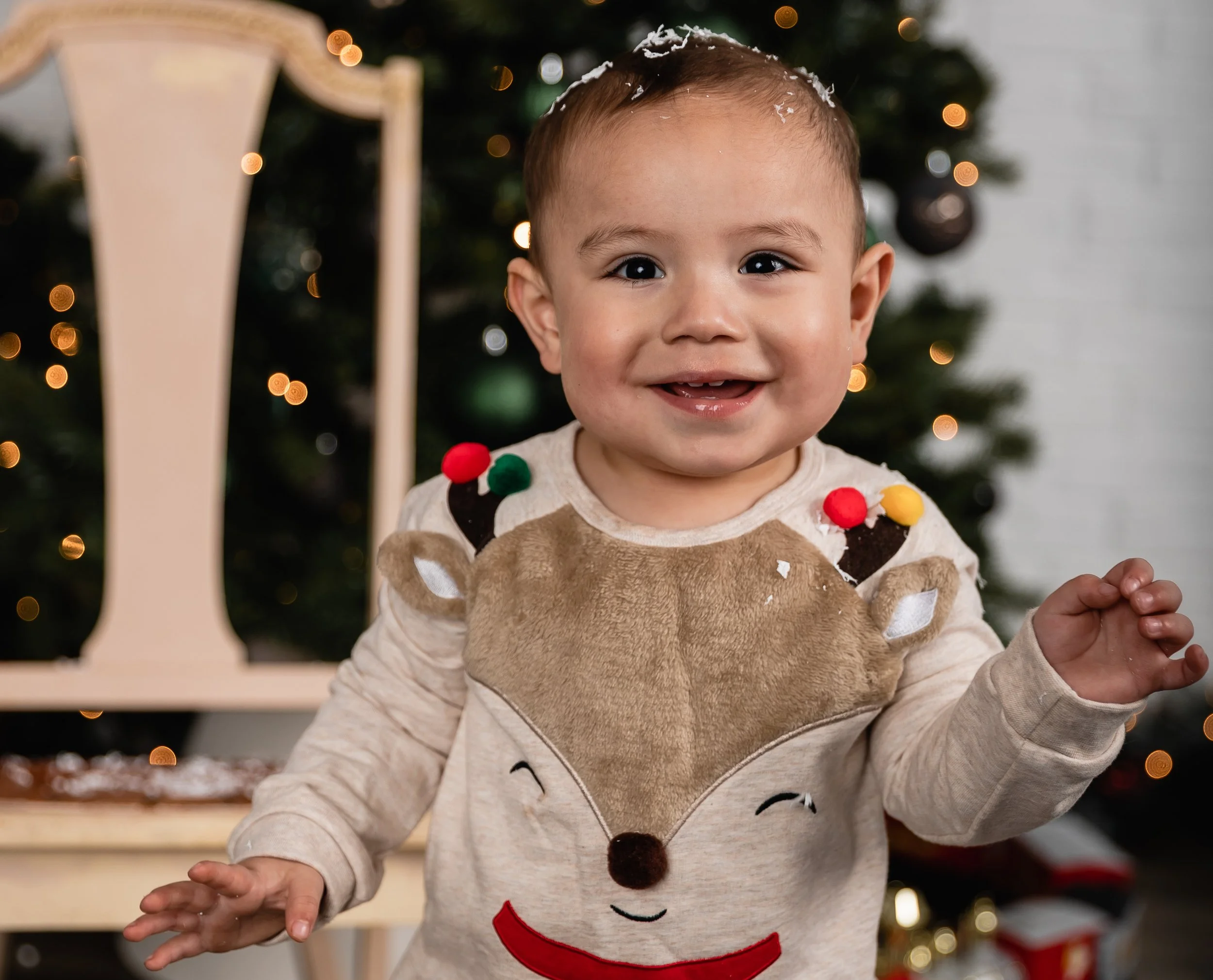 A smiling toddler wearing a reindeer-themed Christmas sweater in front of a decorated Christmas tree.
